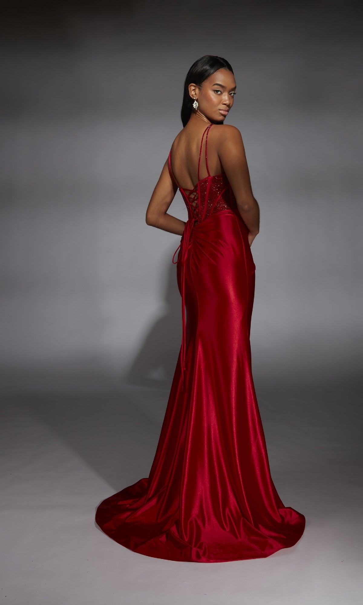 Woman wearing the Alyce 61832 long prom dress with a red satin finish, fitted corset bodice, and lace-up back, standing against a gray studio background and glancing over her shoulder.