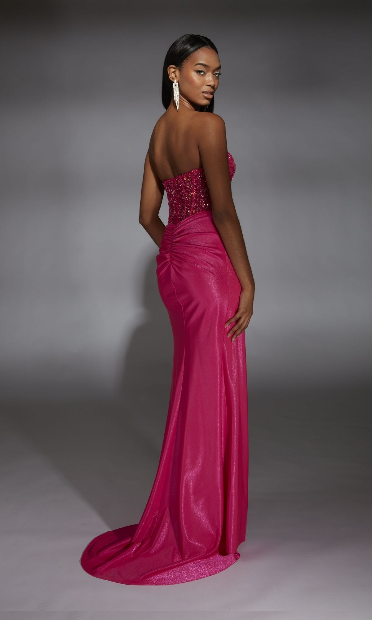 A woman models the Alyce 61844 long prom dress, featuring a hot pink strapless design with an embellished corset bodice, ruched back, and floor-length skirt, posed before a gray backdrop.