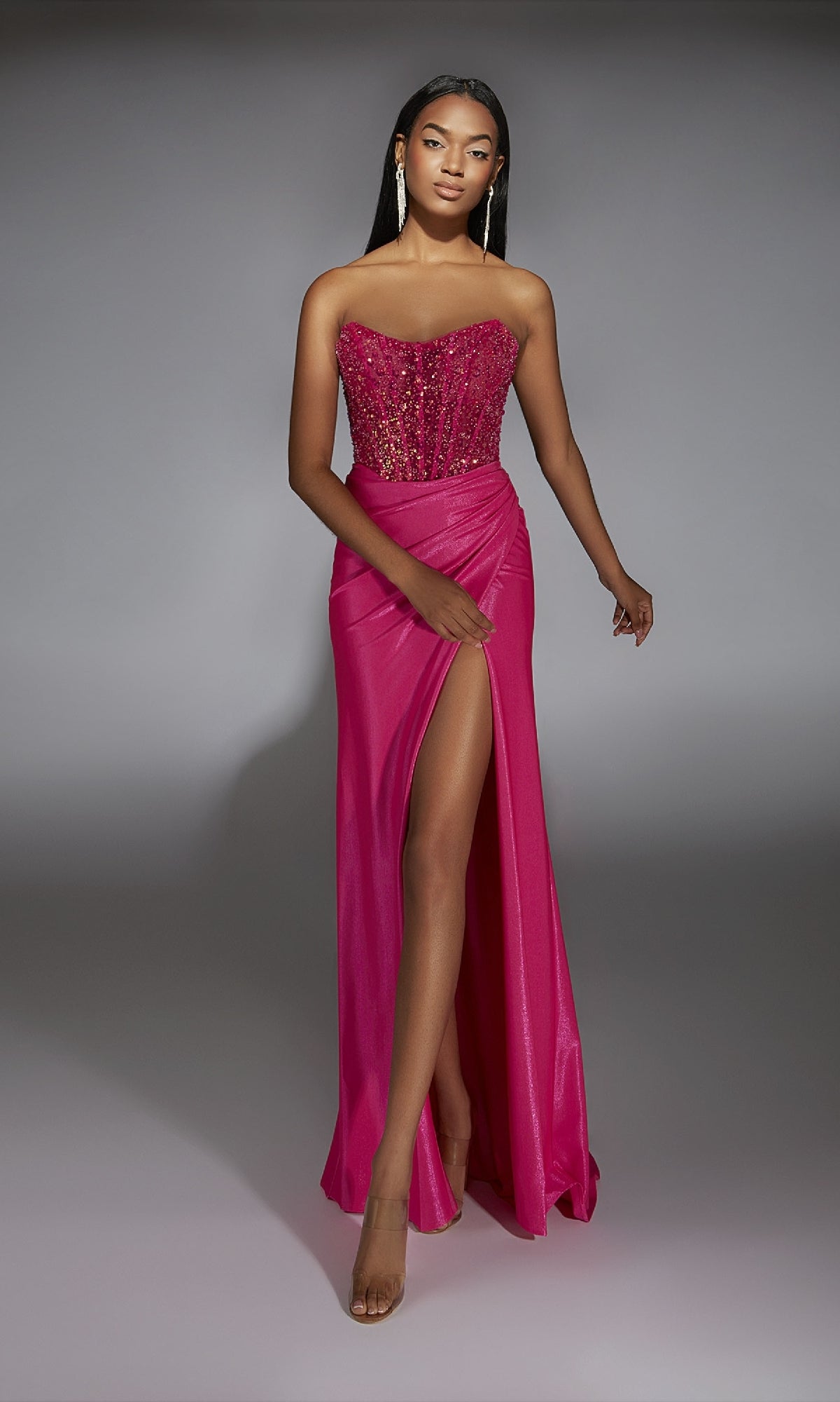 A woman poses against a gray background wearing the Alyce 61844 long prom dress in bright pink, with a strapless embellished corset bodice and high slit.