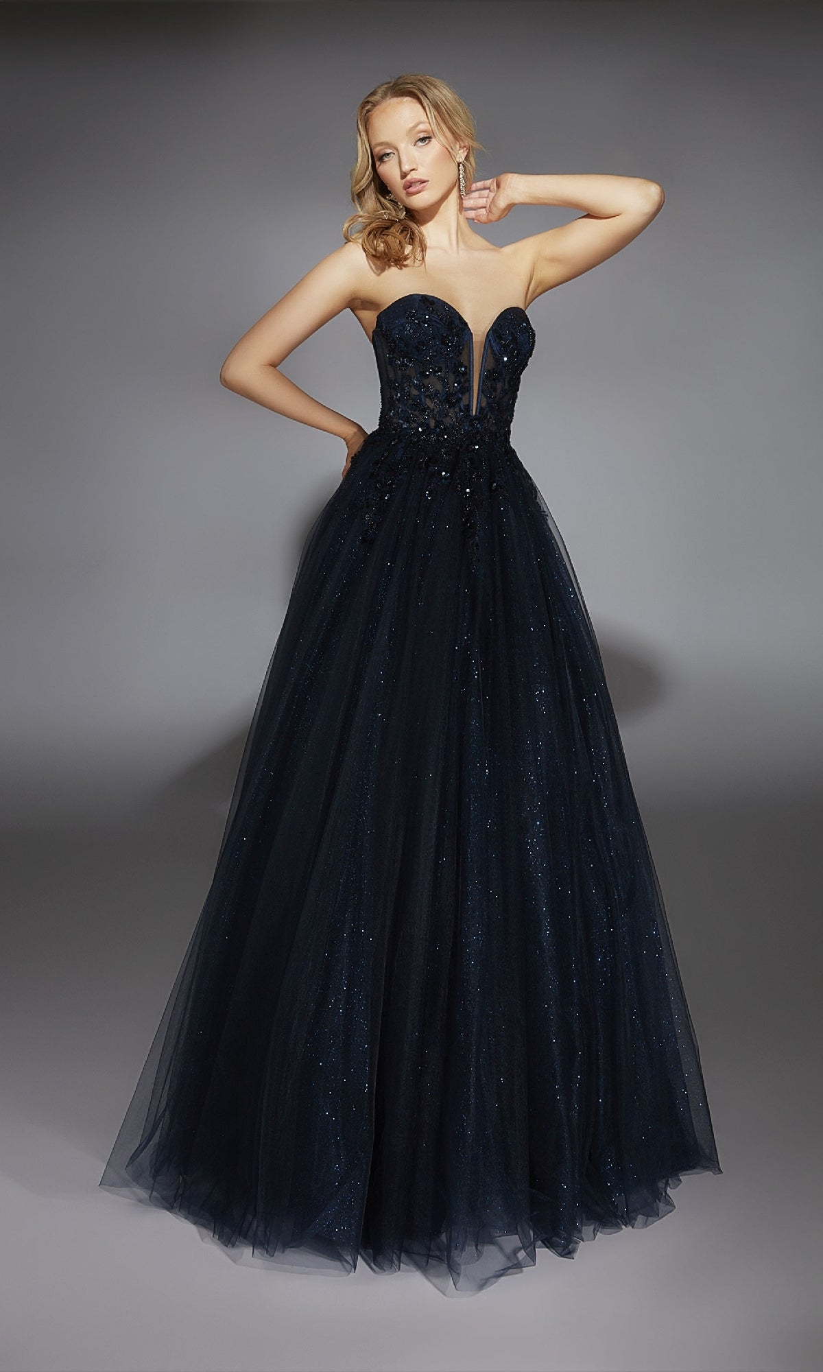 A person wears the Alyce 61846 Strapless Long Glitter Prom Ball Gown, featuring a deep neckline and a full tulle skirt, standing against a plain gray background.