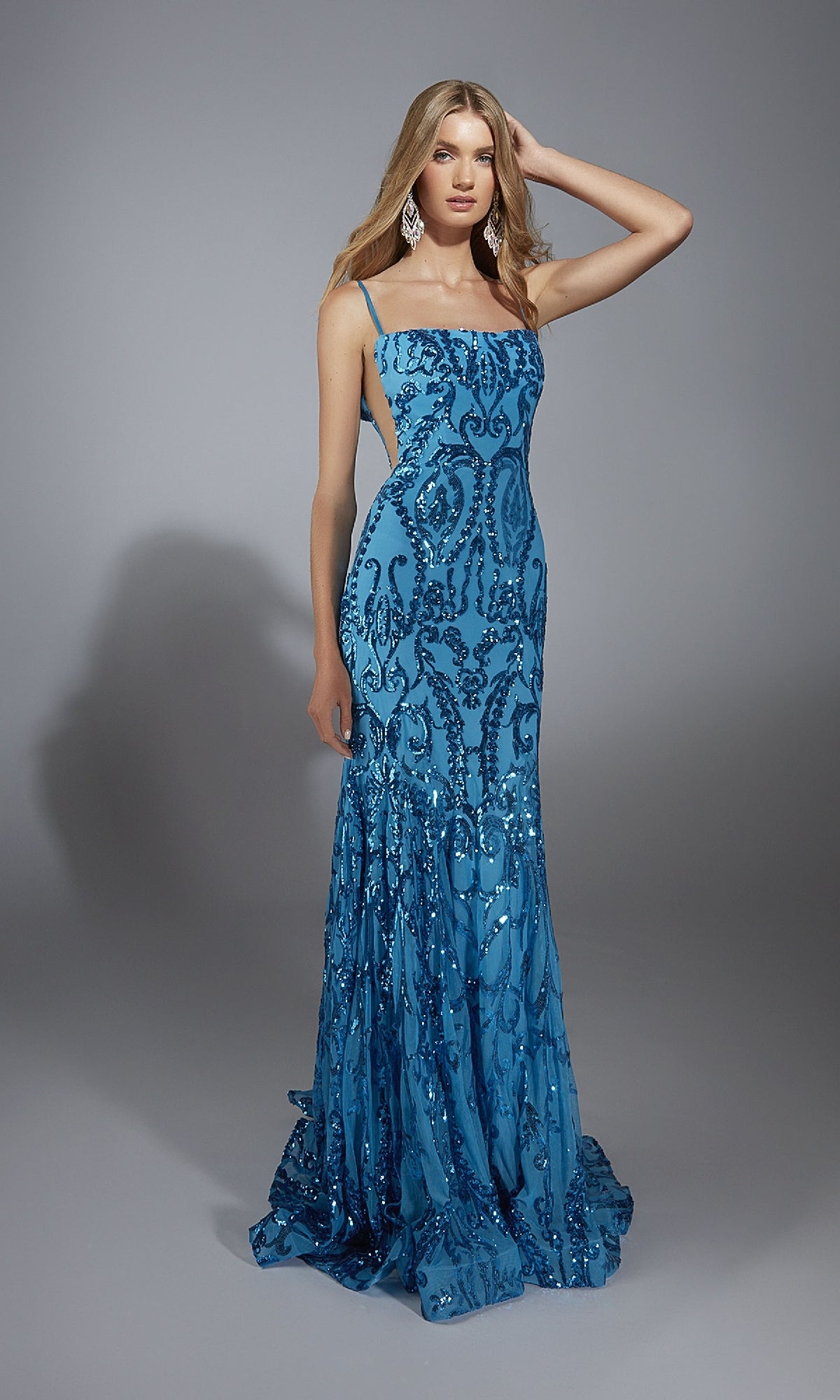 A woman models the Alyce 61852 long prom dress, featuring a fitted blue design with sequin patterns, thin straps, and open sides, against a plain gray background.