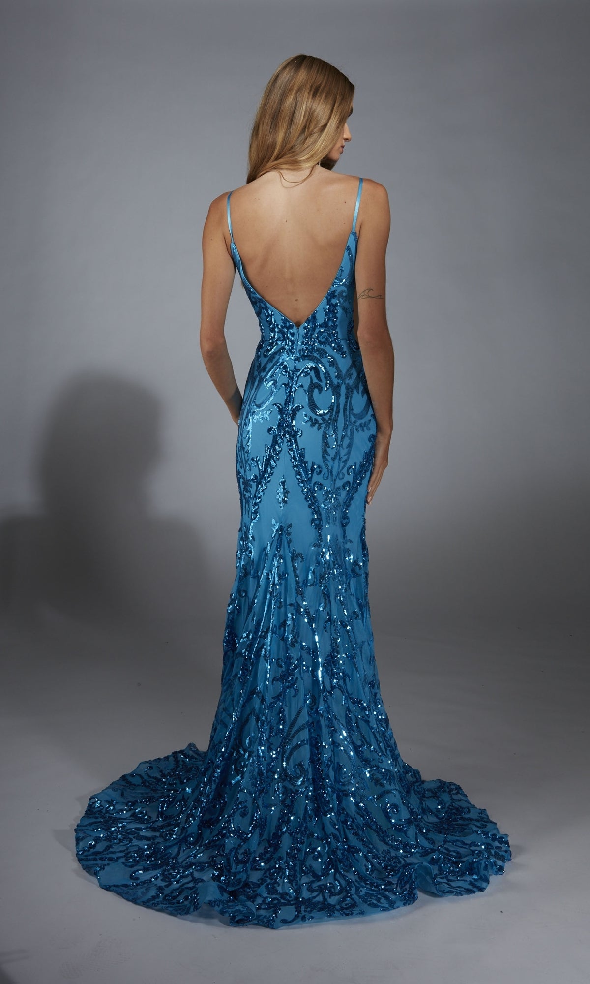 Wearing the Alyce 61852 long prom dress, a woman in a floor-length blue sequin gown with a deep V-back stands facing away against a plain gray background.