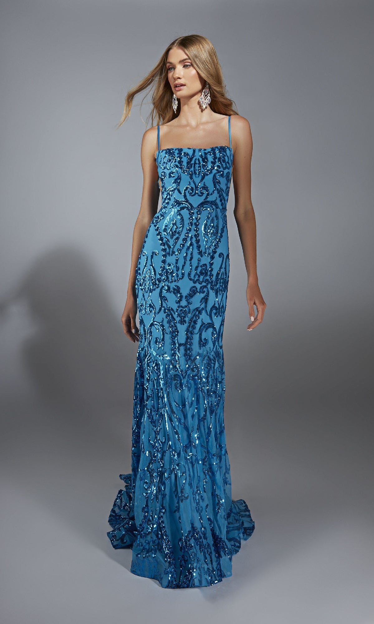 A woman stands against a gray background wearing the Alyce 61852 long prom dress—floor-length, fitted, blue, and embellished with sequins.