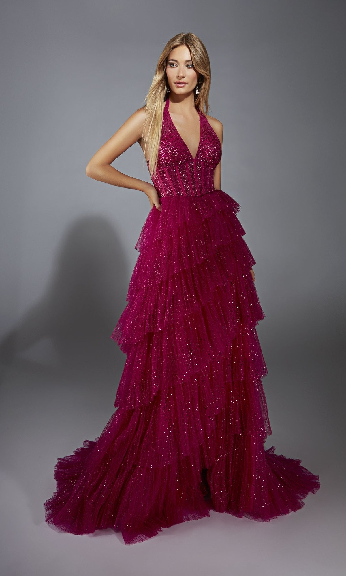 A woman poses in the Alyce 61859 long prom dress, featuring a sleeveless deep pink tiered gown with a corset bodice, deep V-neckline, and glittery accents against a plain gray background.
