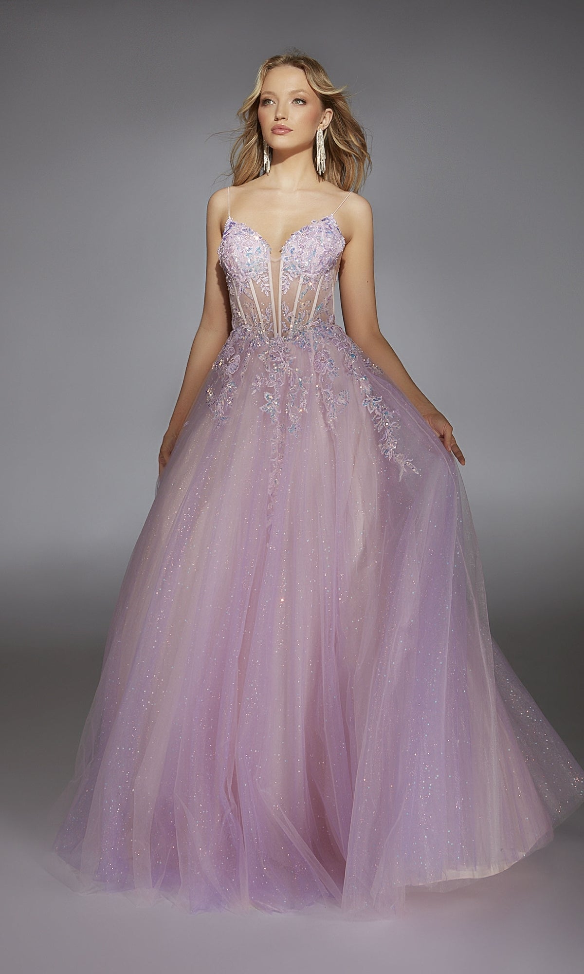 A person models the Alyce 61865 long prom dress, featuring a floor-length lavender glitter tulle skirt, sheer corset bodice with thin straps, floral appliqué, and lace embroidery, standing against a gray background.