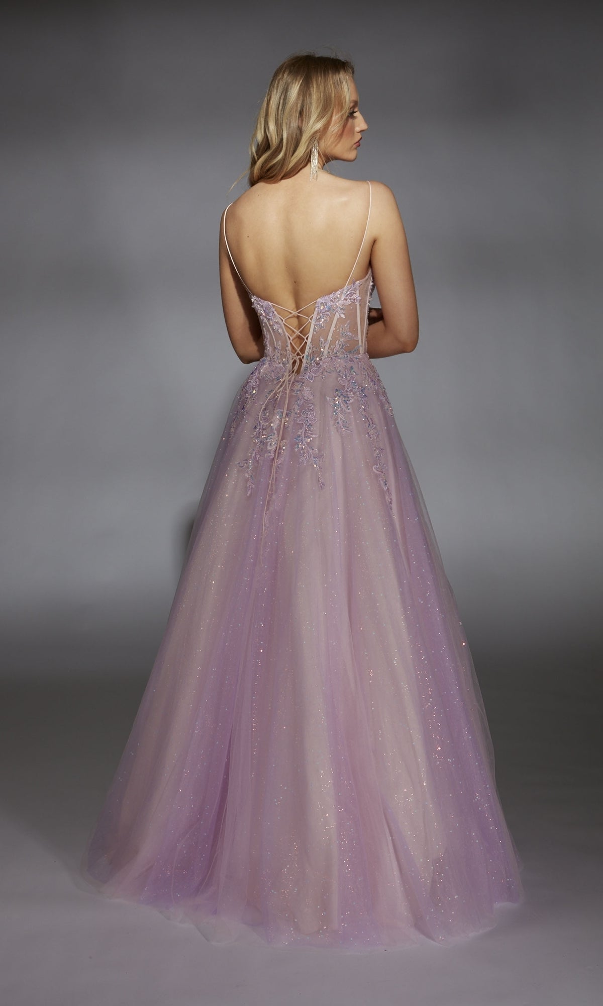 A woman in the Alyce 61865 long prom dress, featuring a lavender hue, floor-length backless design, thin straps, lace-up detailing, and a corset bodice that accentuates her graceful silhouette, stands facing away from the camera.