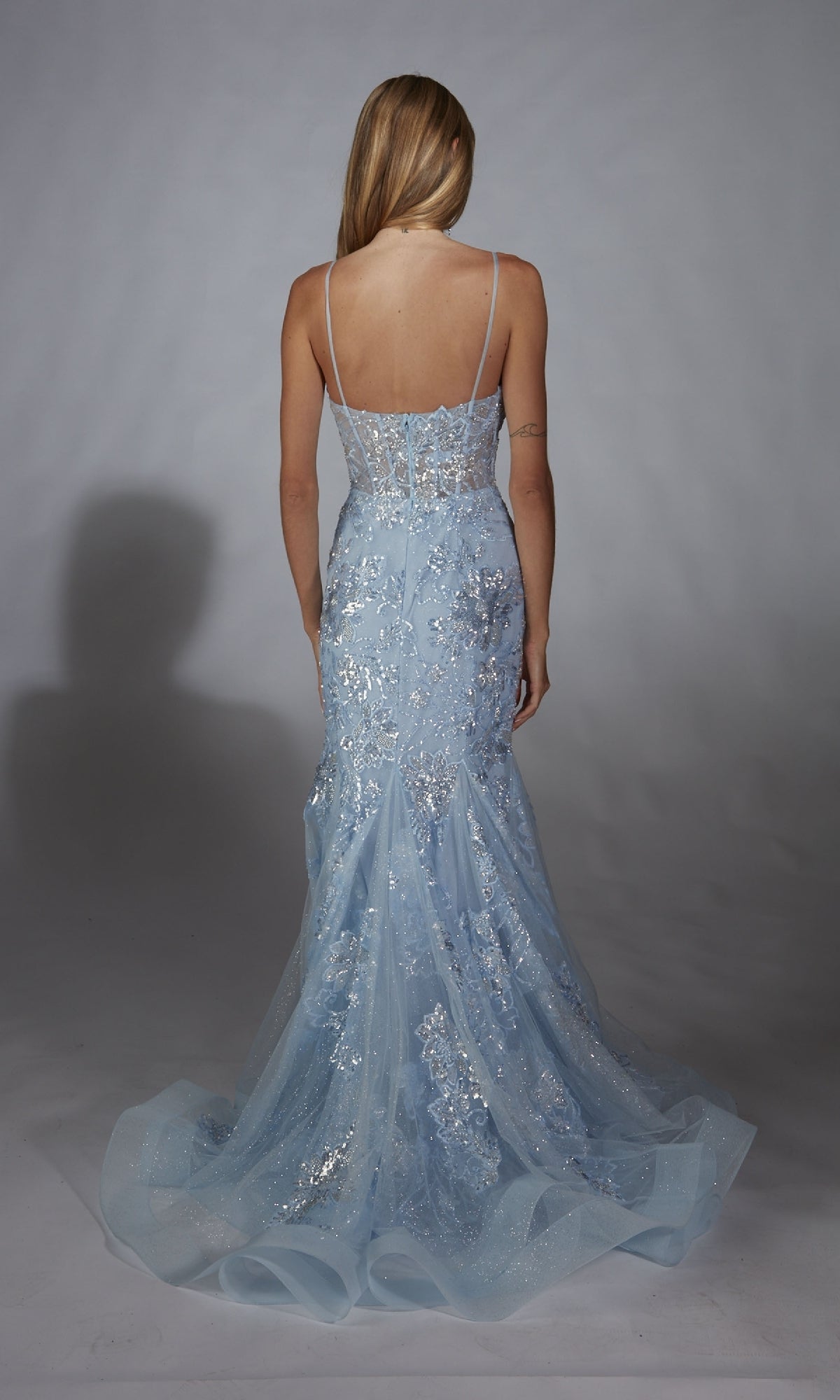 A woman faces away in the Alyce 61867 Long Prom Dress, a light blue, floor-length gown with a sheer corset bodice, lace accents, sleeveless design, and a flowing train displayed against a plain backdrop.