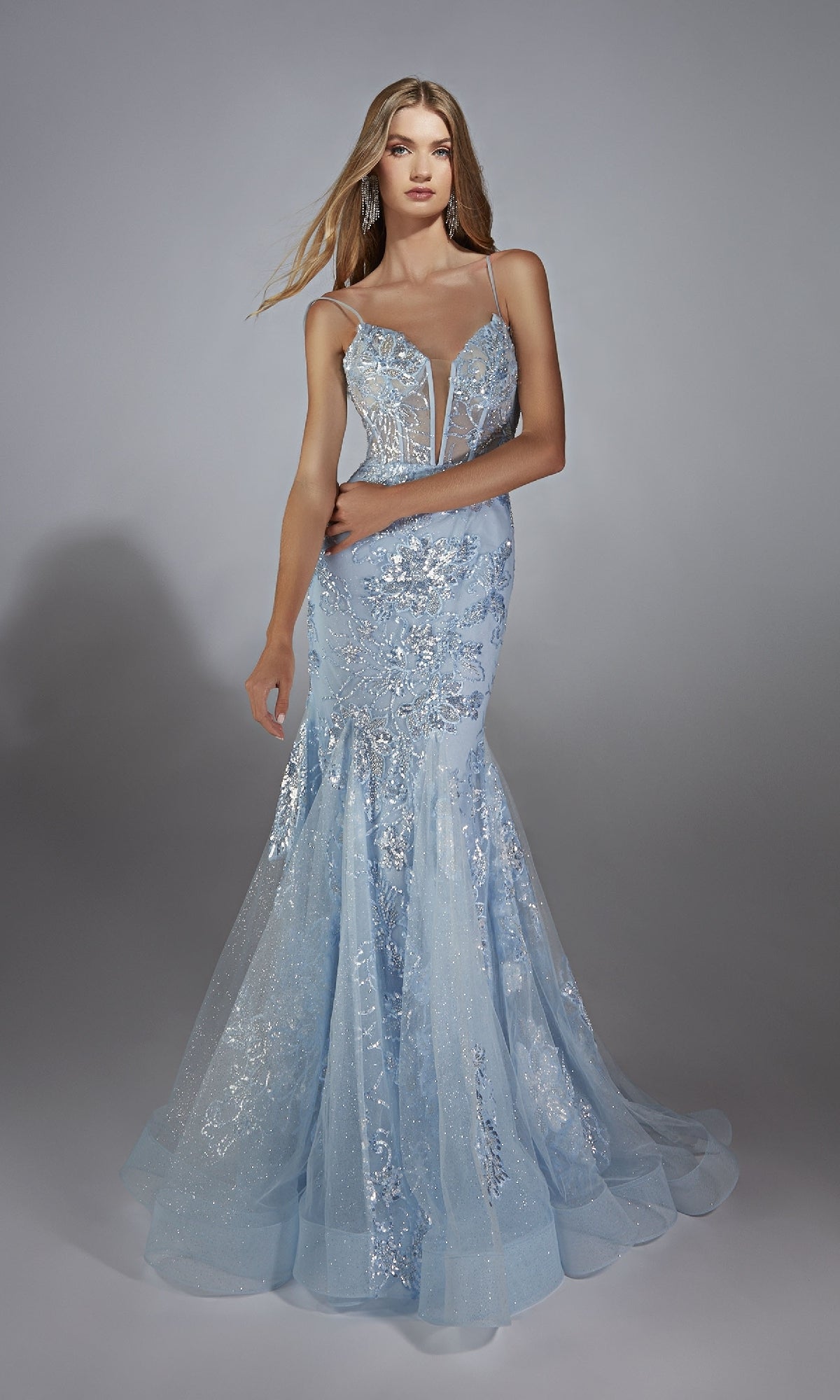 A woman in the Alyce 61867 long prom dress—light blue, floor-length, with sequin embellishments, thin straps, and a tulle mermaid skirt—stands against a plain gray background.