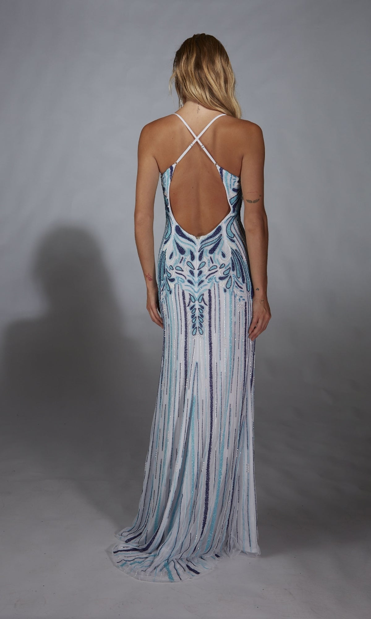 Woman wearing the Long Prom Dress: Alyce 61868, a sleeveless sequin gown with blue and white beaded patterns and a thigh-high slit, stands against a plain gray background facing away from the camera.