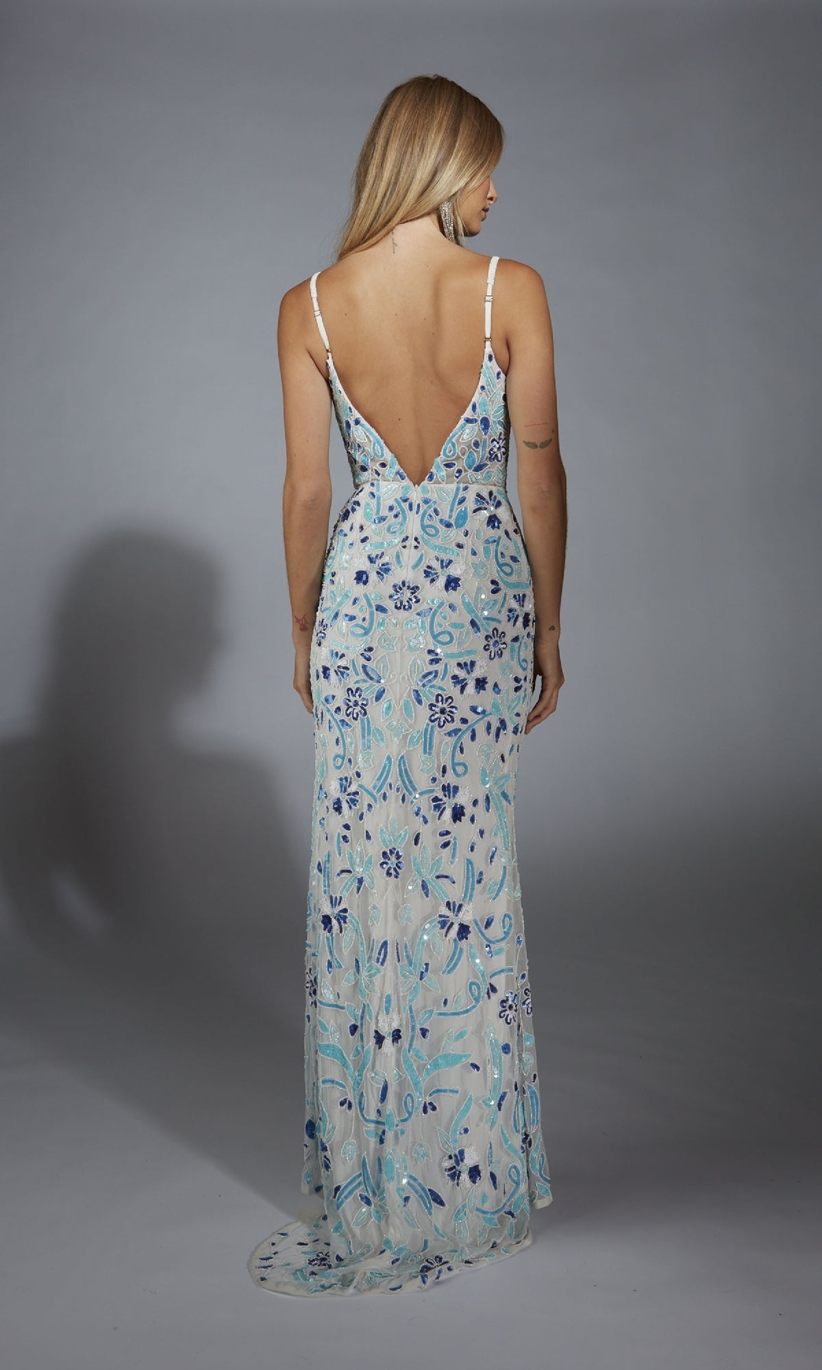 A woman faces away, modeling the Alyce Paris Long Prom Dress 61871—a floor-length, sleeveless gown with blue floral patterns and a deep V-back—set against a plain gray background.