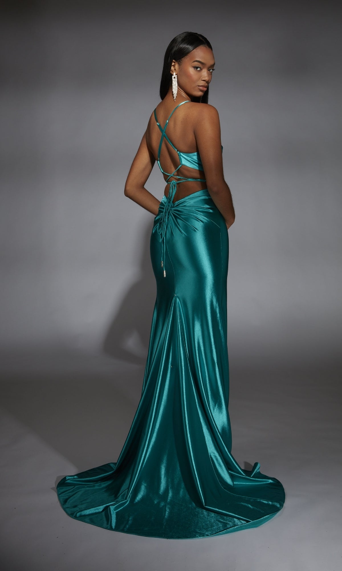 A woman poses against a gray background in the Alyce 61875 long prom dress, featuring teal satin fabric, a backless crisscross strap design, and a floor-length train as she looks over her shoulder.