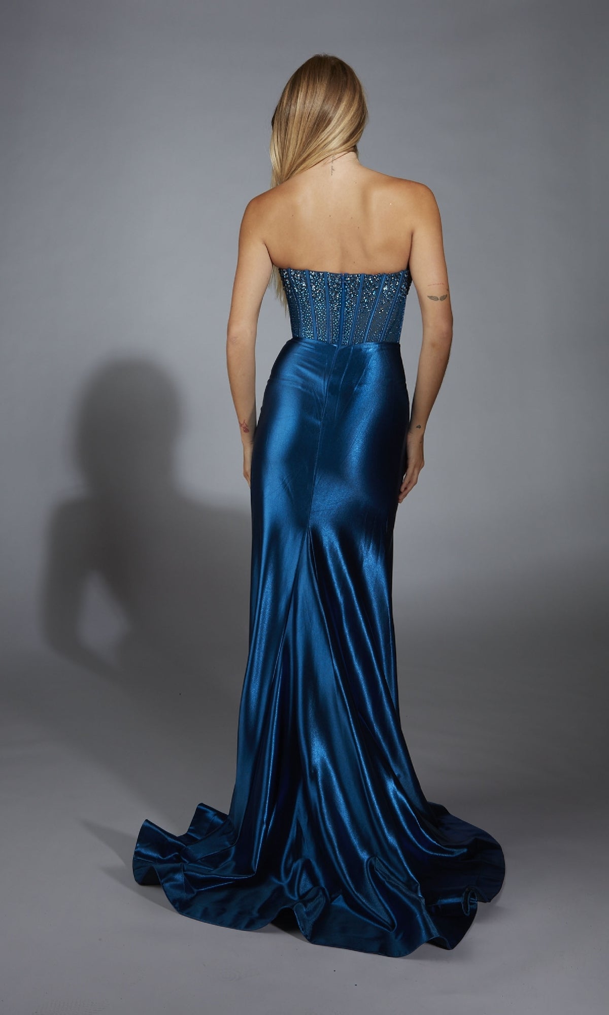 A woman with long blonde hair stands with her back to the camera in front of a gray background, wearing the Alyce 61879 strapless long satin prom dress with a beaded corset-style bodice.