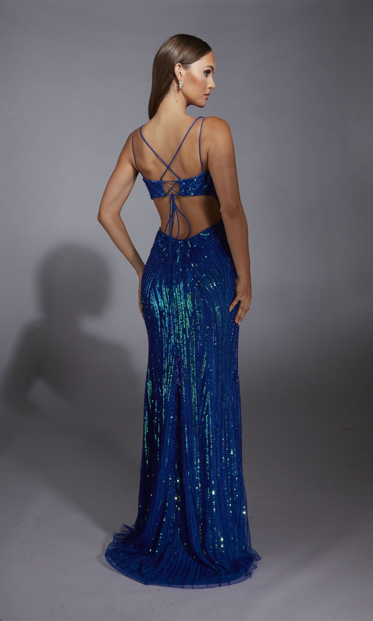 A woman faces away, modeling the Alyce 61880 long prom dress—floor-length with blue sequins, thin straps, and an open back—set against a plain gray background.