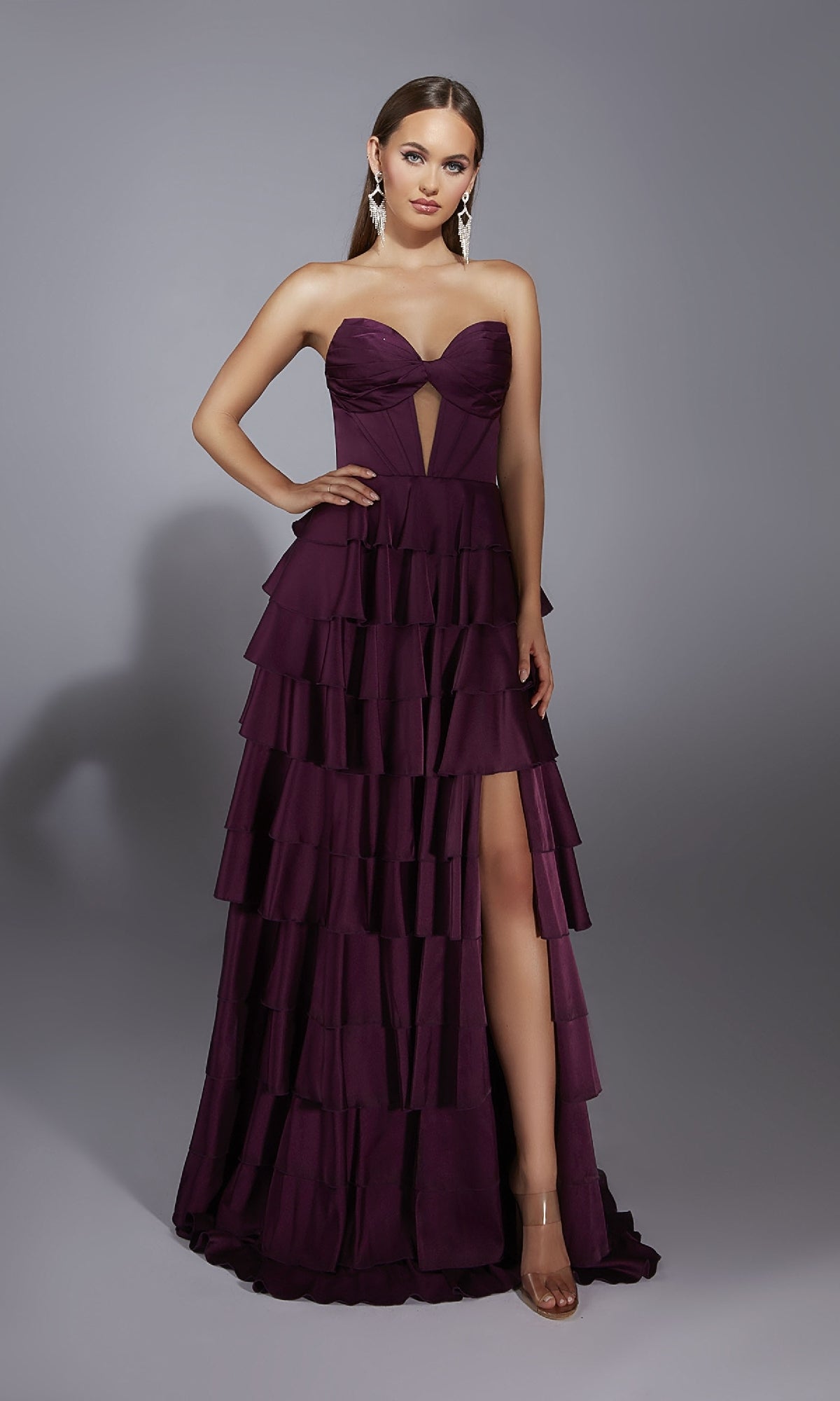 Woman wearing the Long Prom Dress: Alyce 61889, a strapless gown with a deep purple ruffled A-line tiered skirt and front slit, paired with clear heels and long earrings, stands against a plain gray background.
