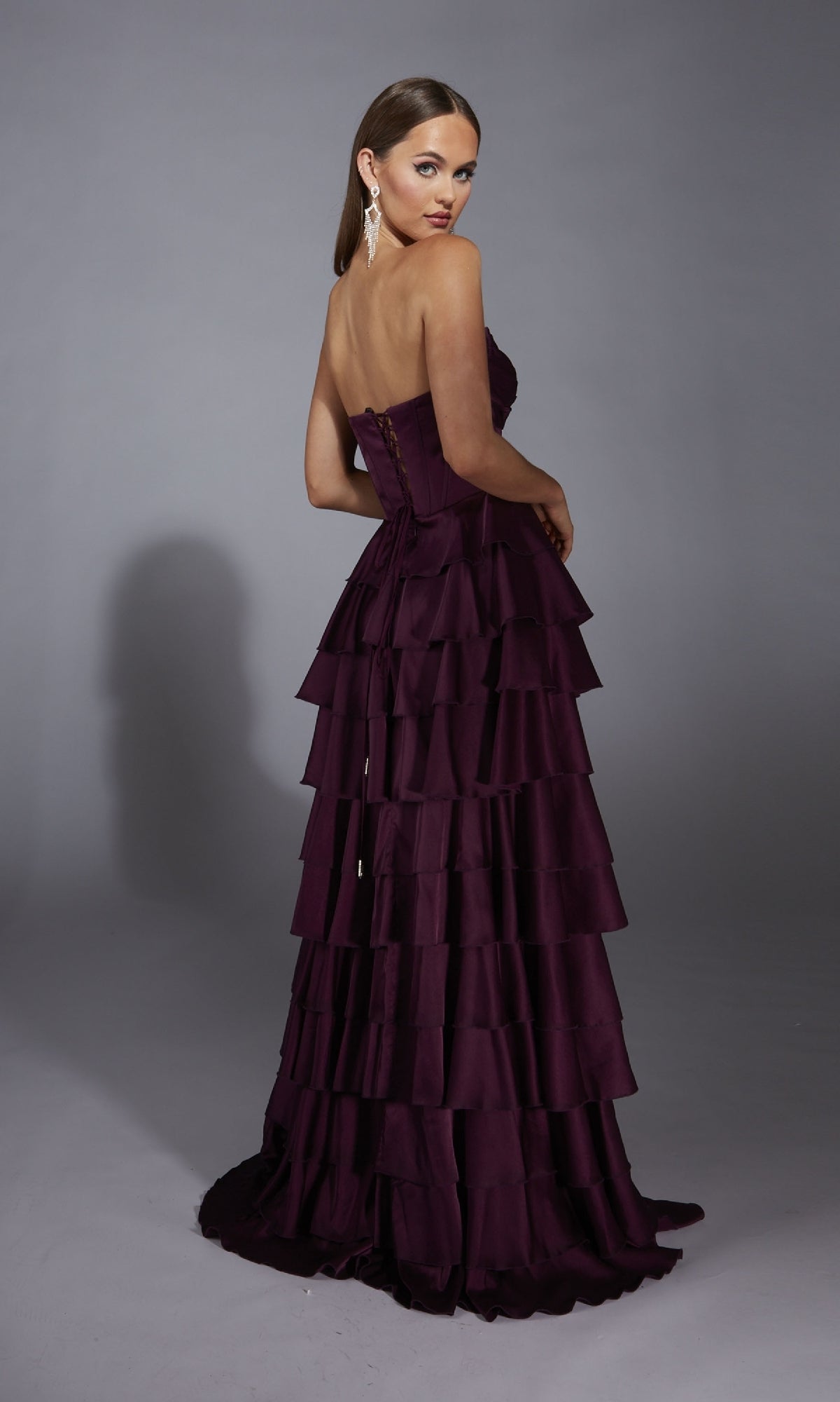 A woman poses against a plain gray background in the Alyce 61889, a strapless long prom dress with an A-line tiered skirt, looking over her shoulder.