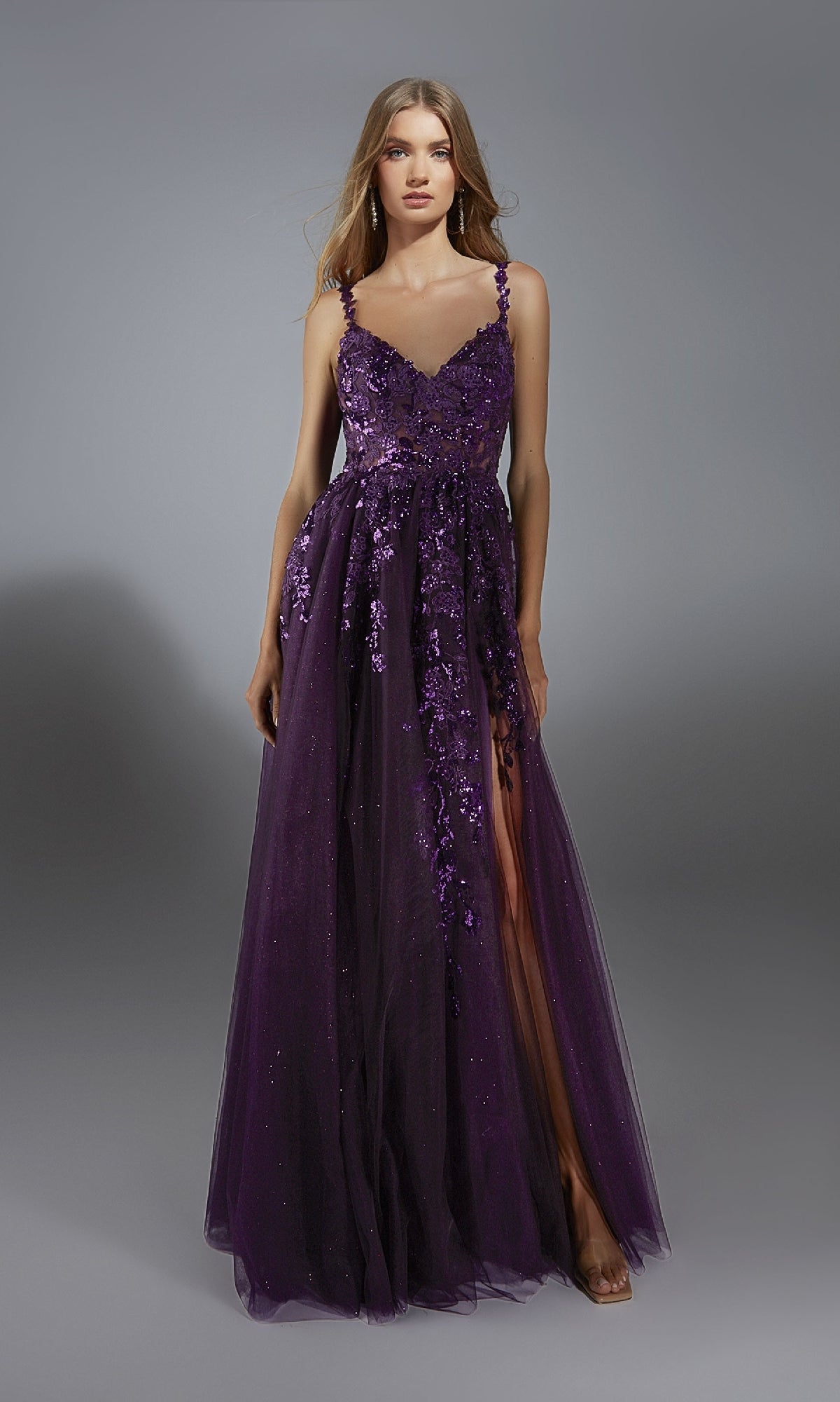 A woman stands against a gray background in the Alyce 61891 long prom dress—a sleeveless, floor-length dark purple gown featuring floral embellishments, lace appliques, and an A-line glitter tulle skirt.