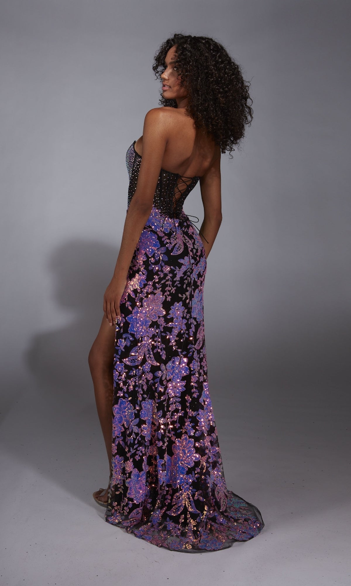 A woman poses in a studio wearing the Alyce 61898 Long Prom Dress, featuring a strapless purple and pink sequin floral print, beaded corset bodice, floor-length skirt with side slit, and looks over her shoulder.