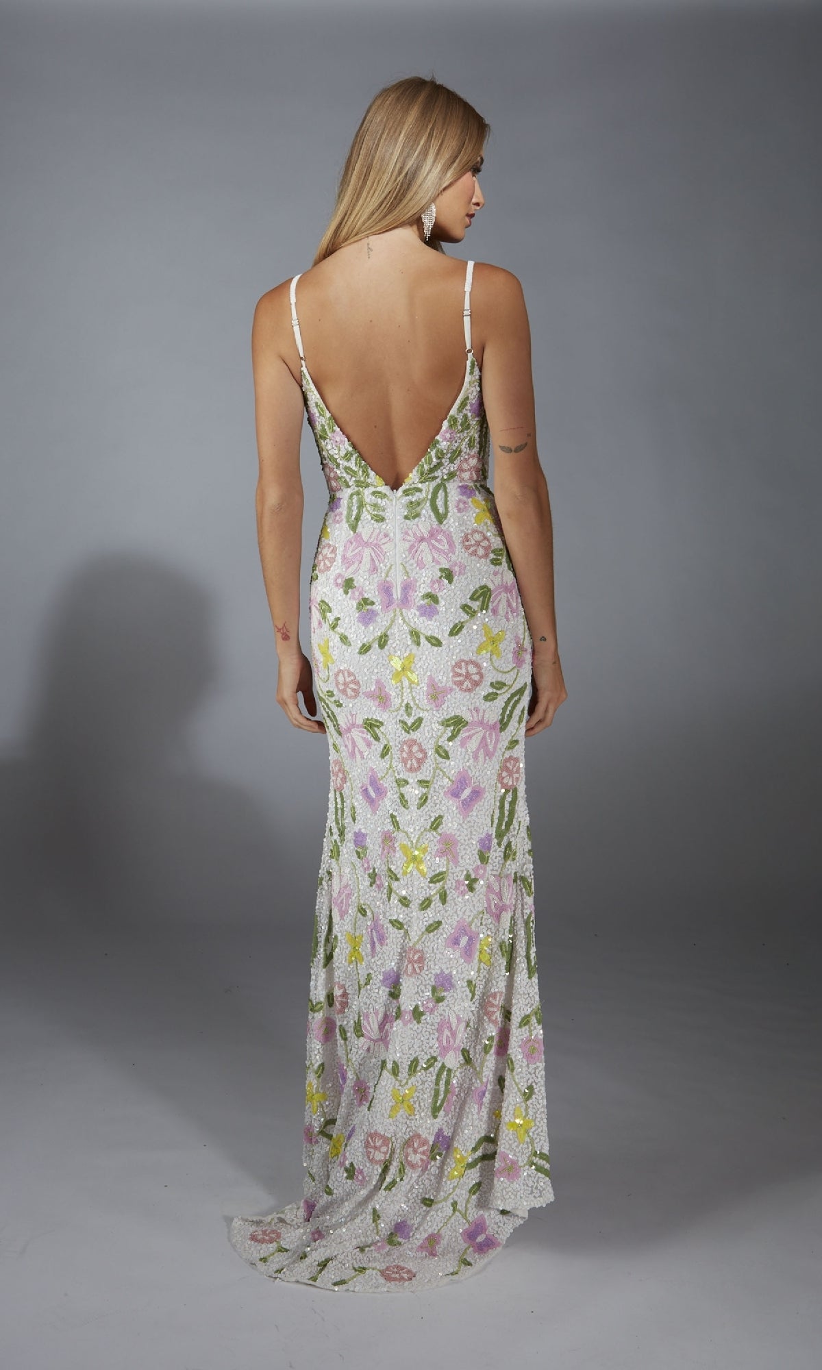 A person with long blonde hair wears the Alyce 61905 long prom dress, a floral sequin gown with adjustable spaghetti straps and a deep V-back, standing against a plain gray background.