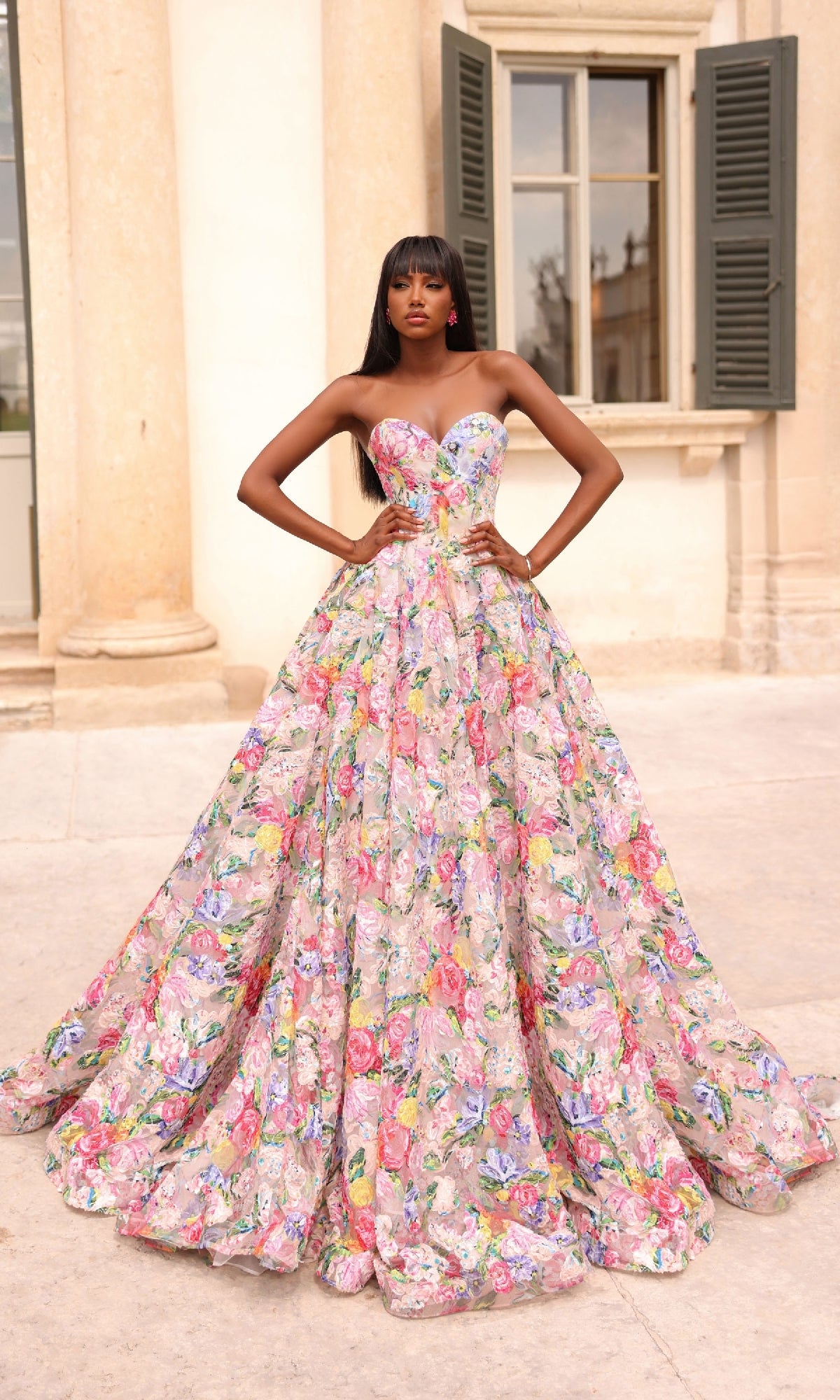 A woman stands outside in front of a building, wearing the Alyce 61913 long prom dress with a strapless bodice and a full, floral-printed skirt, hands on hips.
