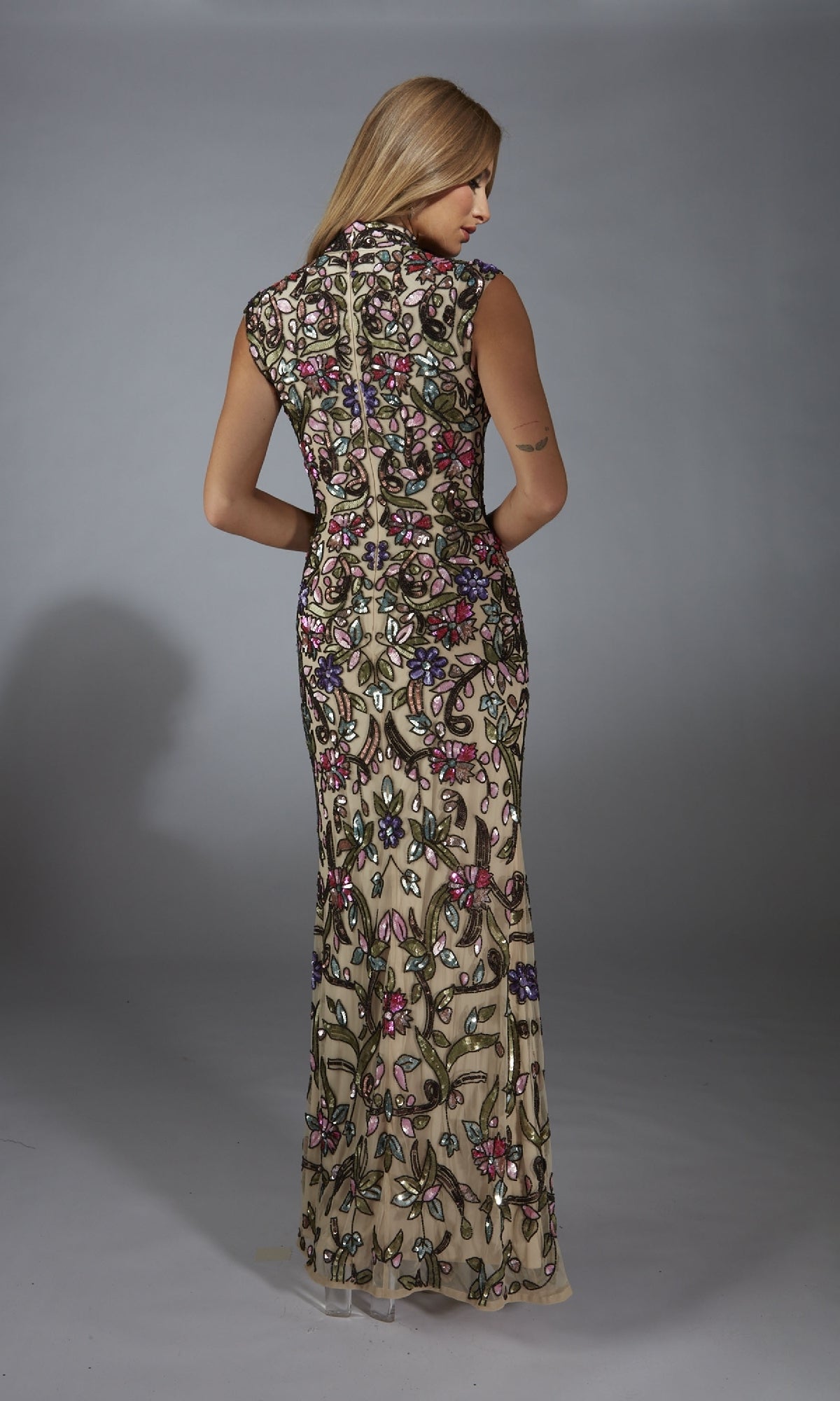 A woman with long blonde hair faces away, wearing the Alyce 61918 Long Prom Dress—a floor-length, sleeveless gown with colorful floral sequin embroidery on sheer nude fabric.
