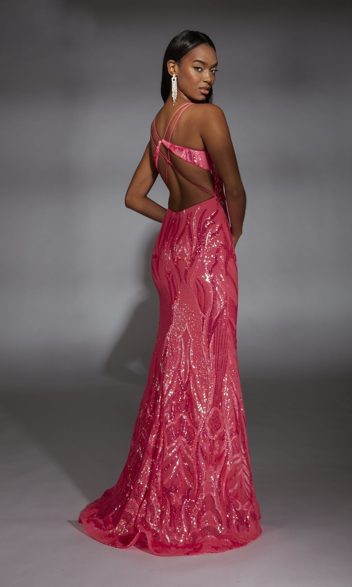 A woman models the Alyce 61930, a floor-length bright pink sequin prom dress featuring a plunging v-neckline, open back, and crisscross straps, as she stands against a neutral gray background.