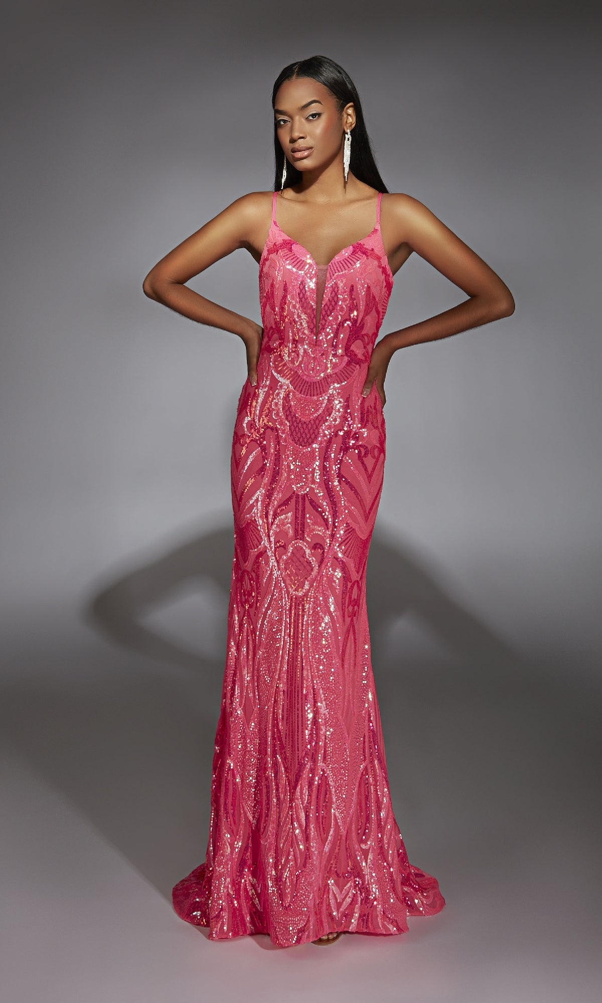 A woman poses with hands on hips in a fitted, floor-length Alyce 61930 sequin prom dress with a plunging neckline, set against a plain gray background.
