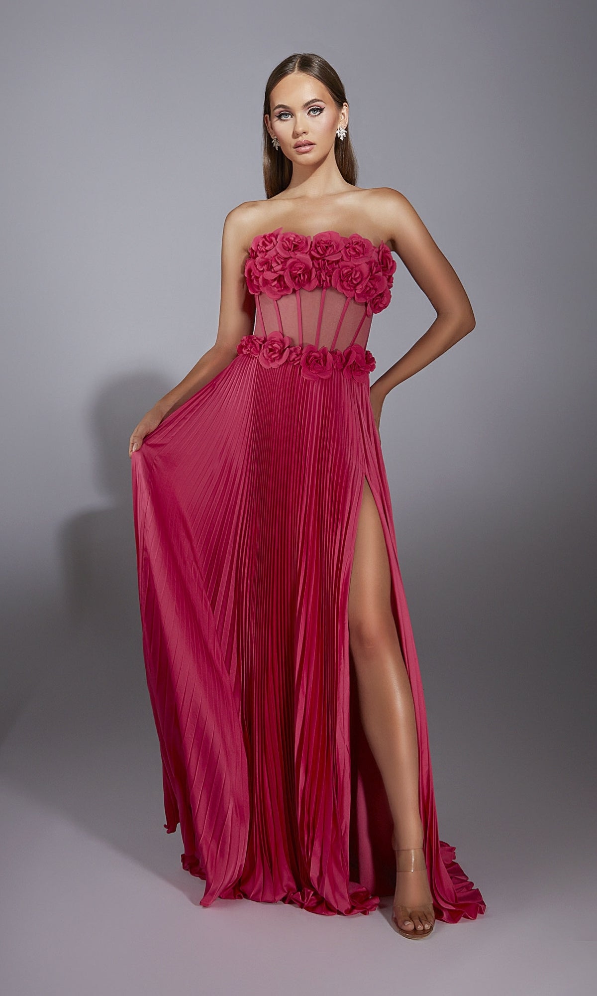 A woman models the Alyce 61935 long prom dress—magenta, strapless, floor-length—with a sheer corset bodice featuring 3D flower details and a high skirt slit, standing against a plain gray background.