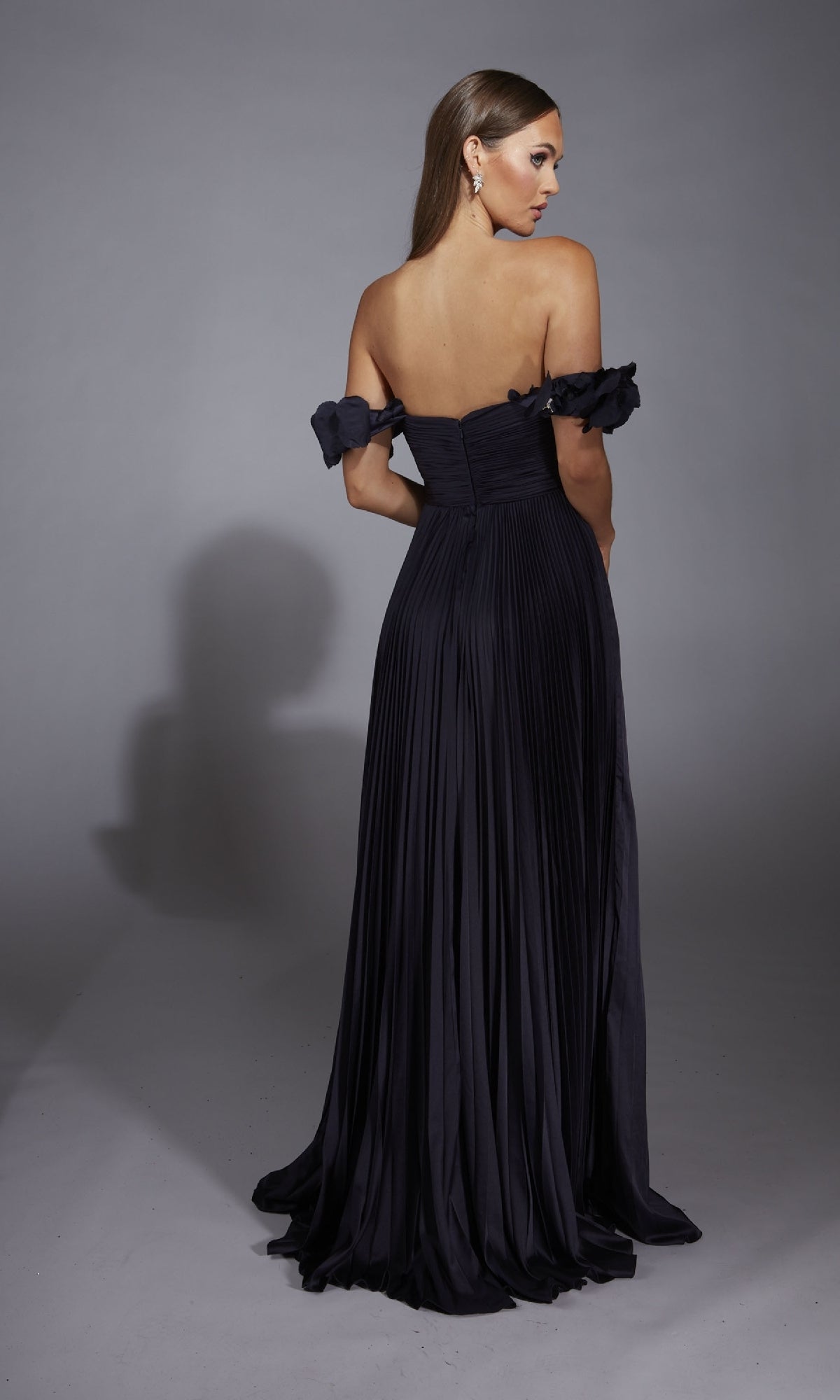A woman stands facing away in a studio with a gray backdrop, wearing the Alyce 61946 long prom dress—an off-the-shoulder, pleated A-line gown.
