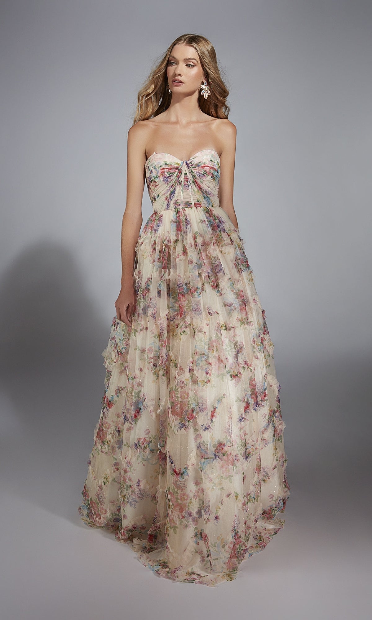 A woman wears the Alyce 61957 long prom dress, featuring a strapless floral design with a corset bodice and flowing A-line skirt, standing against a plain gray background.