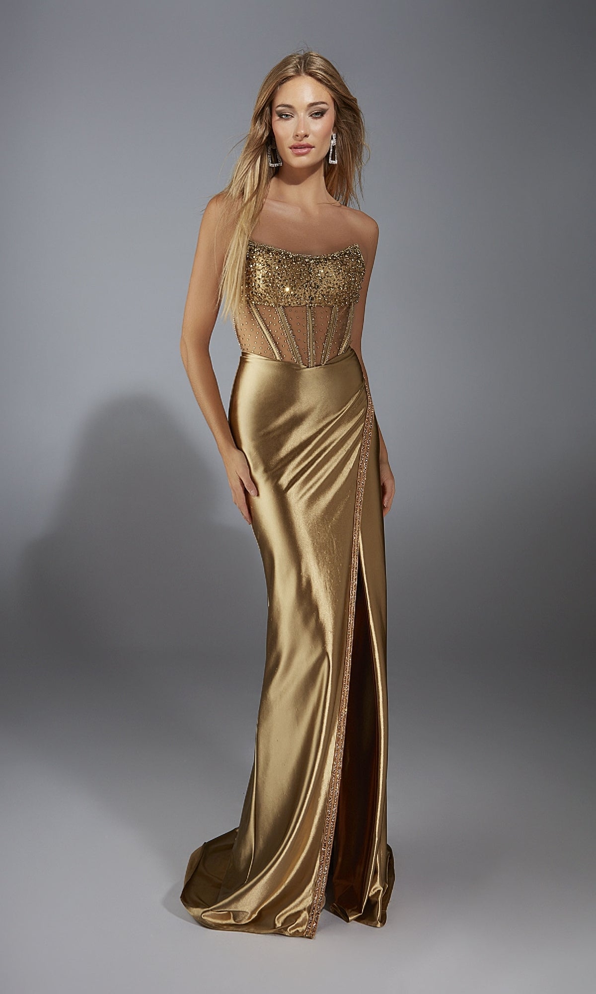 A woman in the Long Prom Dress: Alyce 61976, a gold strapless gown with a sequin corset bodice and thigh-high slit, stands against a plain gray background.
