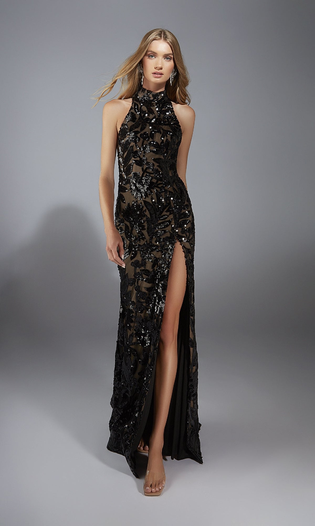 A woman poses against a plain gray background wearing the Alyce 61979 long prom dress, featuring a sleeveless, high-neck design with a high slit and shimmering sequin details.