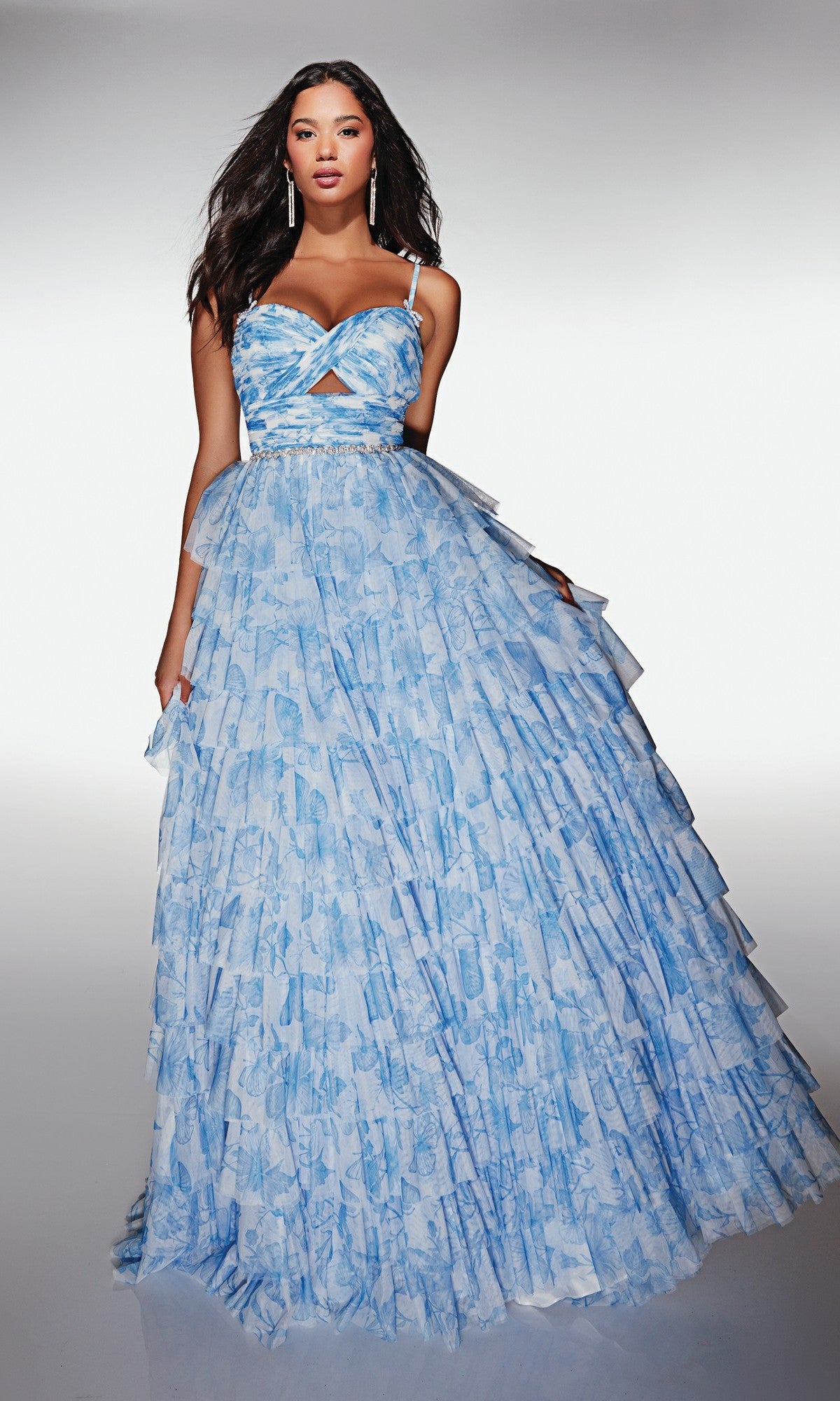 A woman wears the Alyce 62018 long prom dress—light blue, patterned, with thin straps, a waist cutout, and tiered layers—standing against a plain white background.