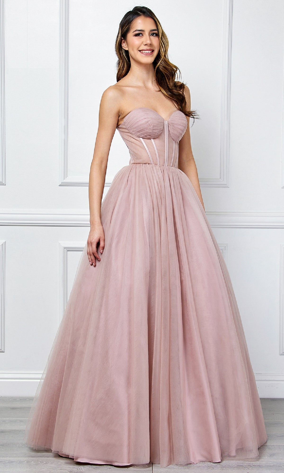A woman models the Amelia Couture 6202 Strapless Corset Prom Ball Gown—a blush pink tulle dress with a fitted bodice and full skirt—standing indoors against a white paneled wall.
