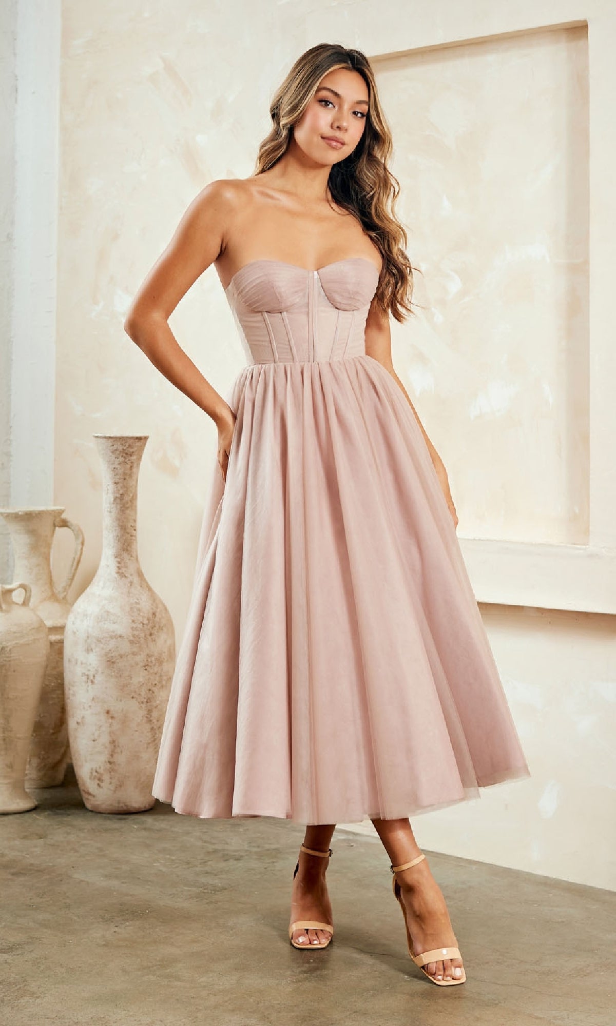 A woman stands indoors in a Strapless Pink Semi-Formal Dress by Amelia Couture with a fitted bodice and flared skirt, paired with nude heels. Large ceramic vases decorate the background.