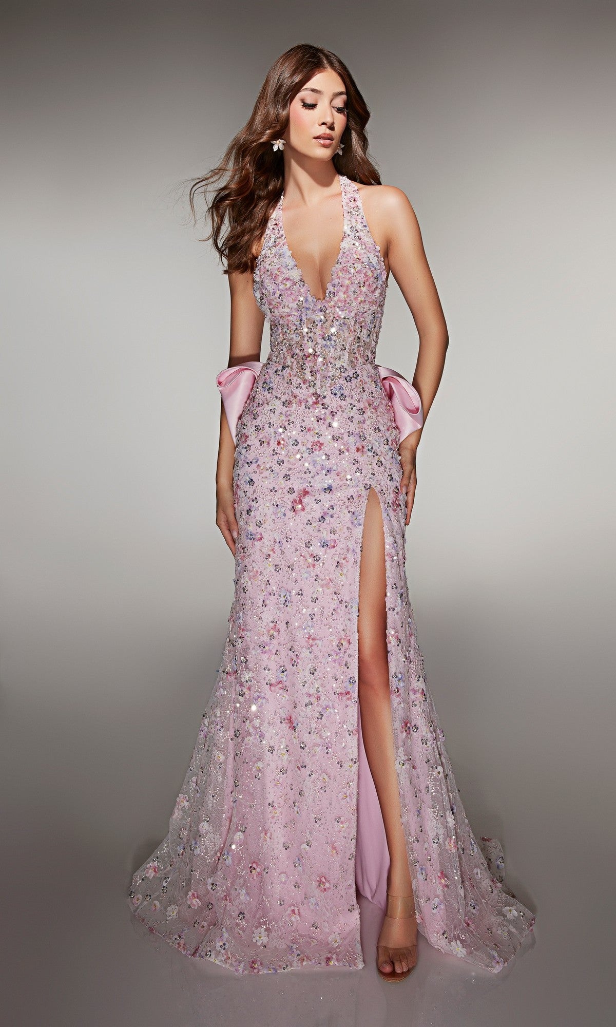 A woman models the Alyce 62044, a long pink prom dress with sequin embellishments, deep neckline, thigh-high slit, and statement satin bow at the back, standing against a plain background.