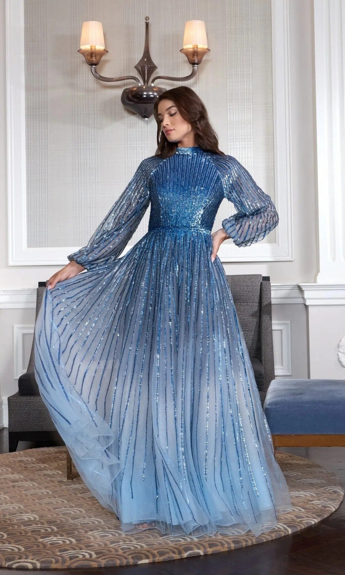A woman stands indoors on a patterned rug beneath wall sconces, wearing the Mac Duggal 6206 long formal dress in slate blue, featuring shimmering ombre sequins and sheer, full sleeves.