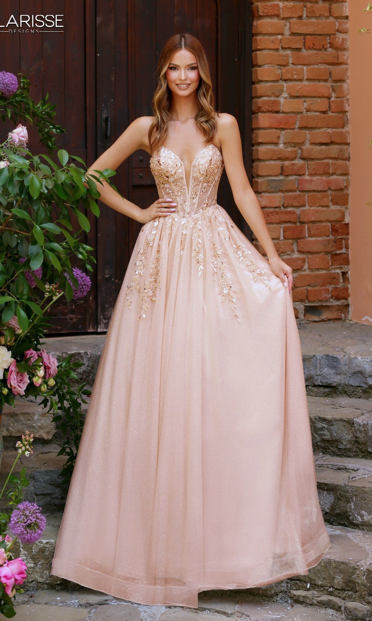 A woman stands outdoors by a brick wall and wooden door, wearing the Clarisse Designs 62102 champagne prom ball gown, featuring beaded floral lace embellishments and a flowing glitter tulle skirt.