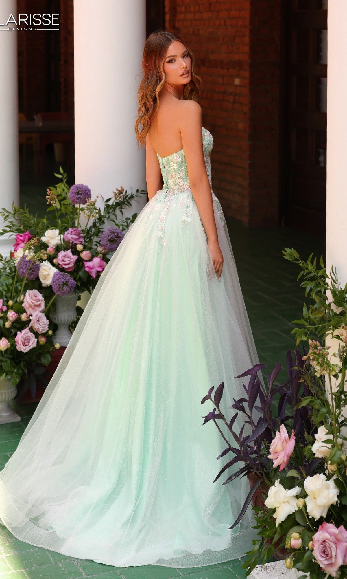 A woman wearing the Clarisse Designs 62135 long prom dress, featuring a strapless corset bodice and floral embroidery, stands among floral arrangements, looking over her shoulder.