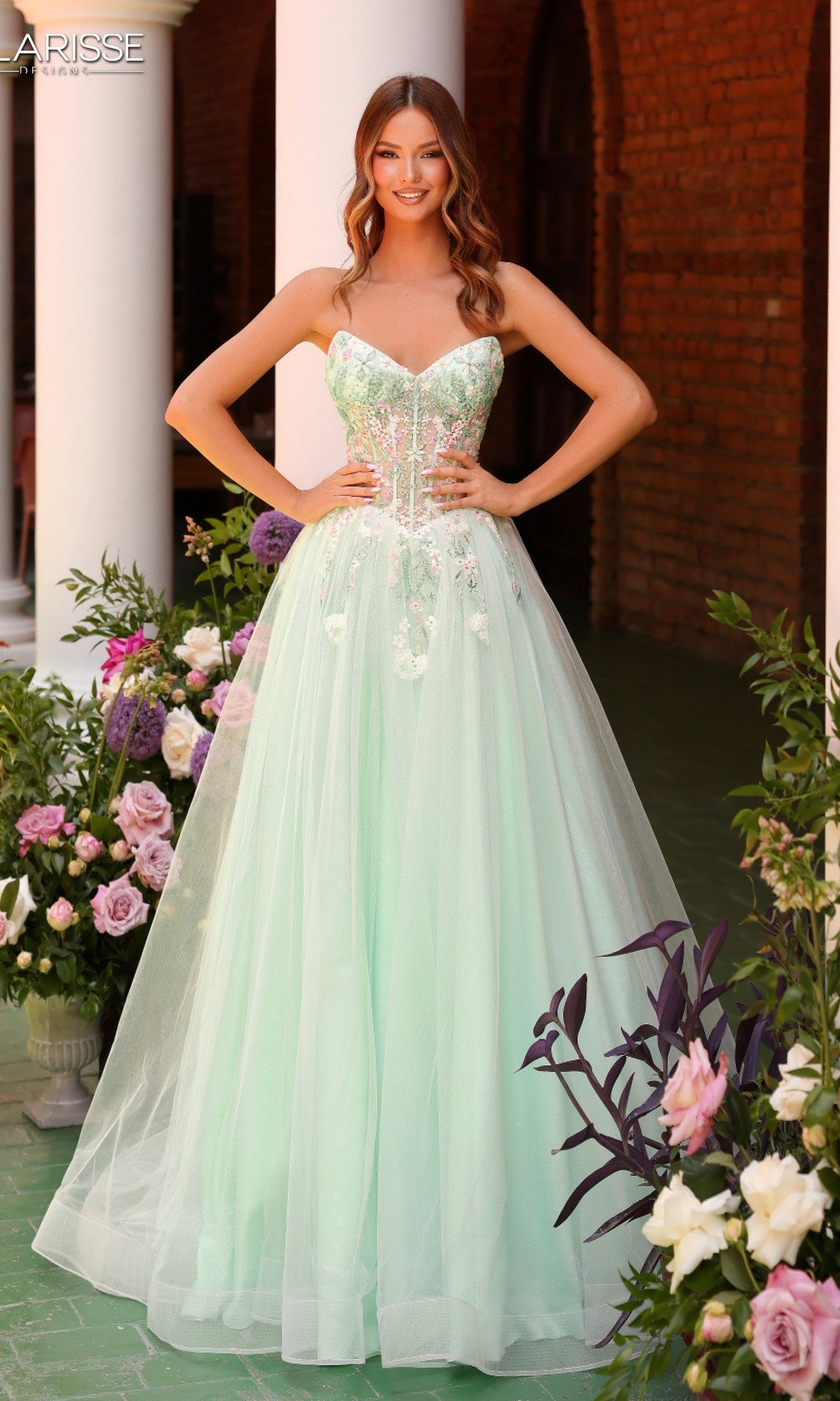 A woman stands outdoors in the Long Prom Dress Clarisse Designs 62135 in seafoam, featuring a strapless corset bodice and delicate floral embroidery, surrounded by beautiful flower arrangements.