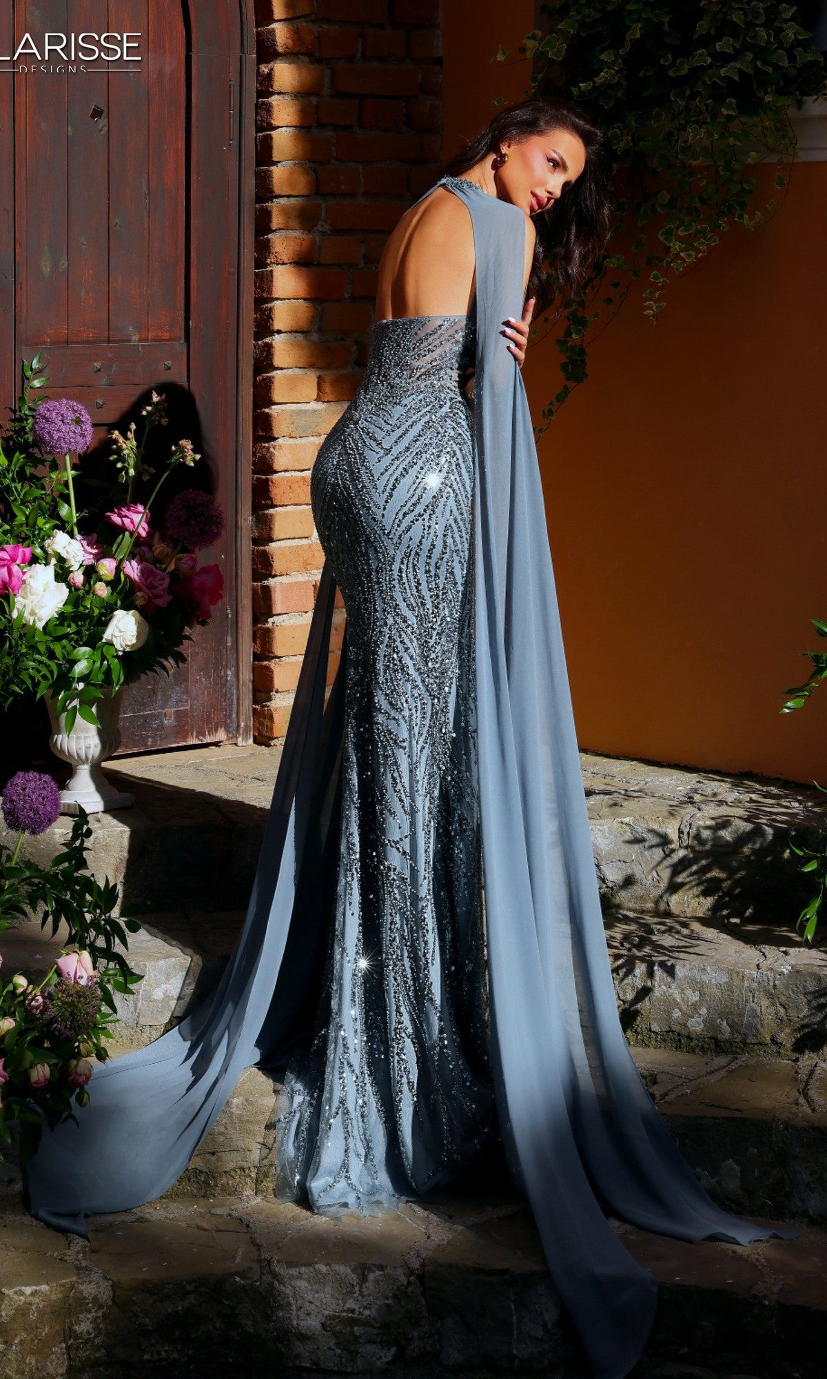 A woman in the Long Formal Dress: Clarisse Designs 62176, a shimmering sequin halter gown with a long train, stands on stone steps near flowers and a wooden door.