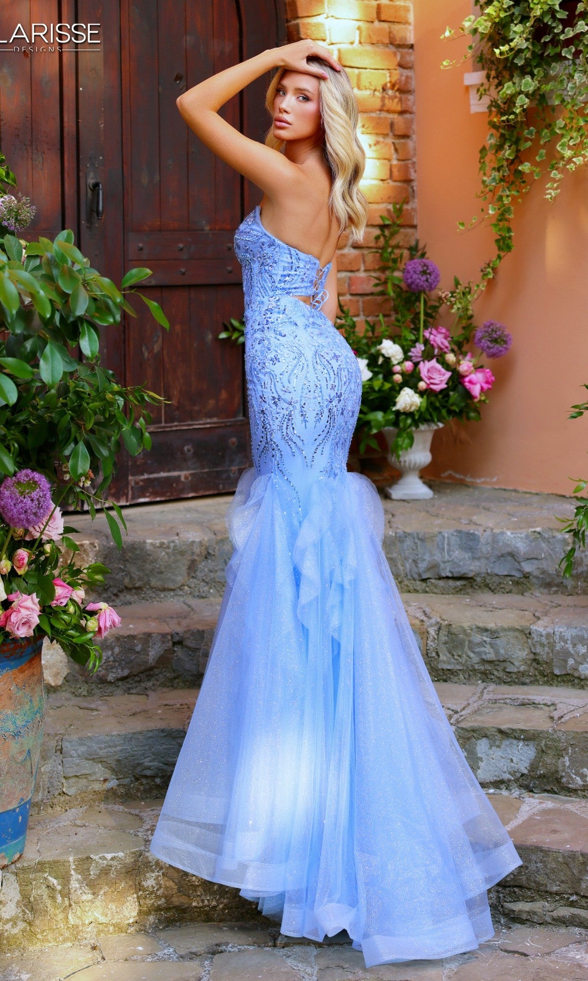 A woman poses outdoors on stone steps near a wooden door and flowers, wearing the Clarisse Designs 62258 long prom dress—a strapless mermaid gown with beaded embroidery.