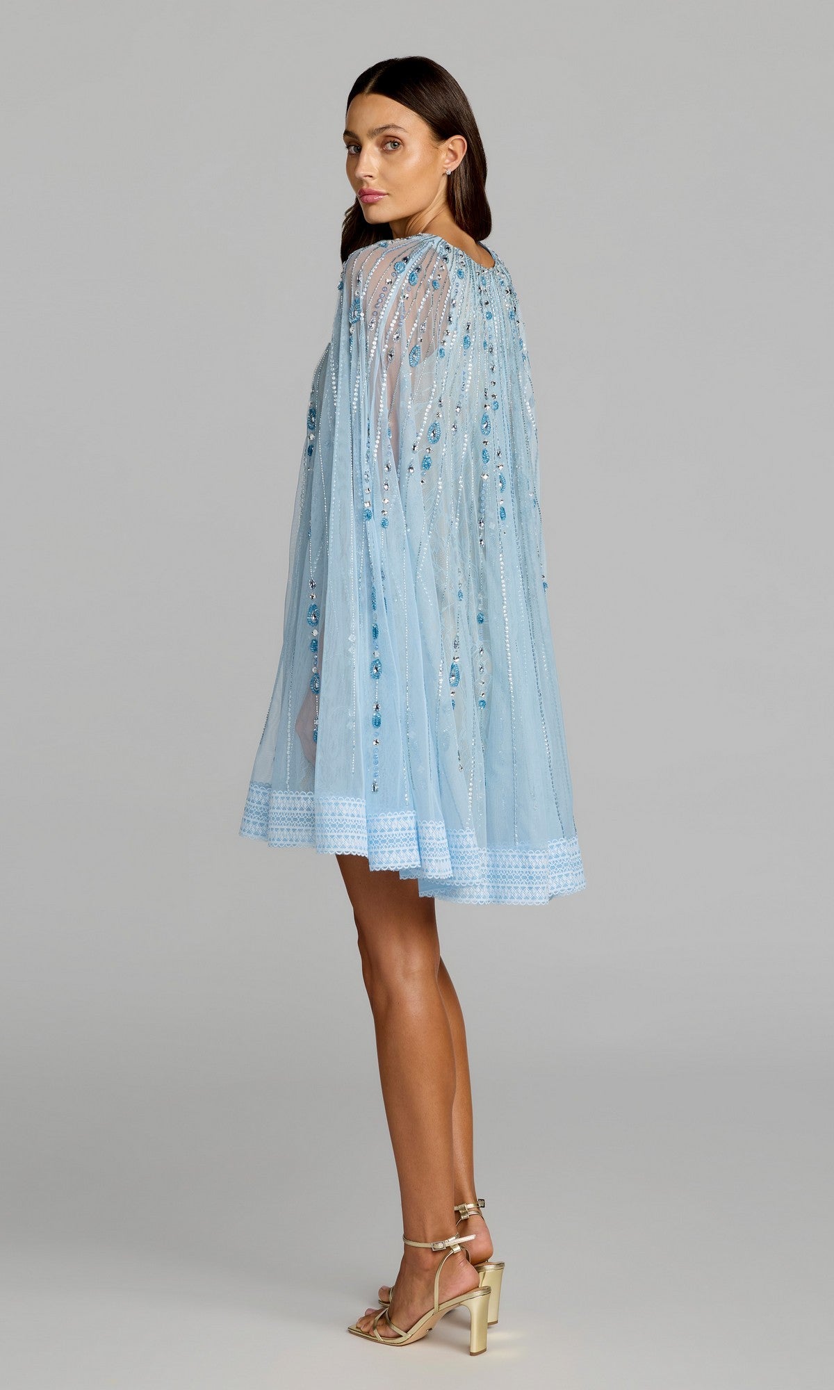 A woman stands in profile against a plain background, wearing the Mac Duggal 6226 short party dress—a sheer, light blue hand-beaded design with cape detail—paired with gold high-heeled sandals.