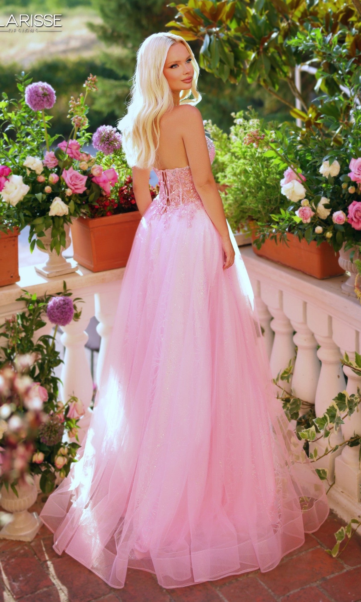 Wearing the Long Prom Dress: Clarisse Designs 62312, a woman in a strapless glitter pink corset gown stands on a balcony among potted flowers, gazing over her shoulder at the camera.