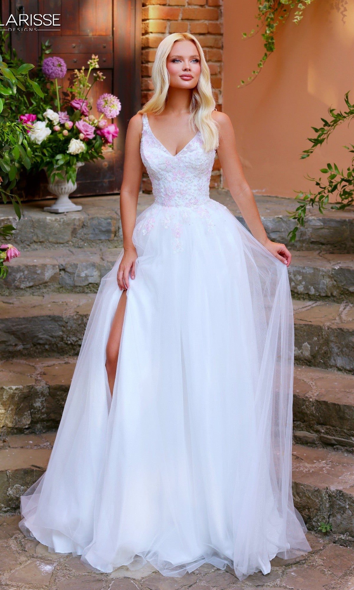 A woman wears the Long Prom Dress: Clarisse Designs 62314 in White/Pink—featuring a beaded embroidered bodice and thigh-high slit—while standing on outdoor stone steps surrounded by greenery and flowers.