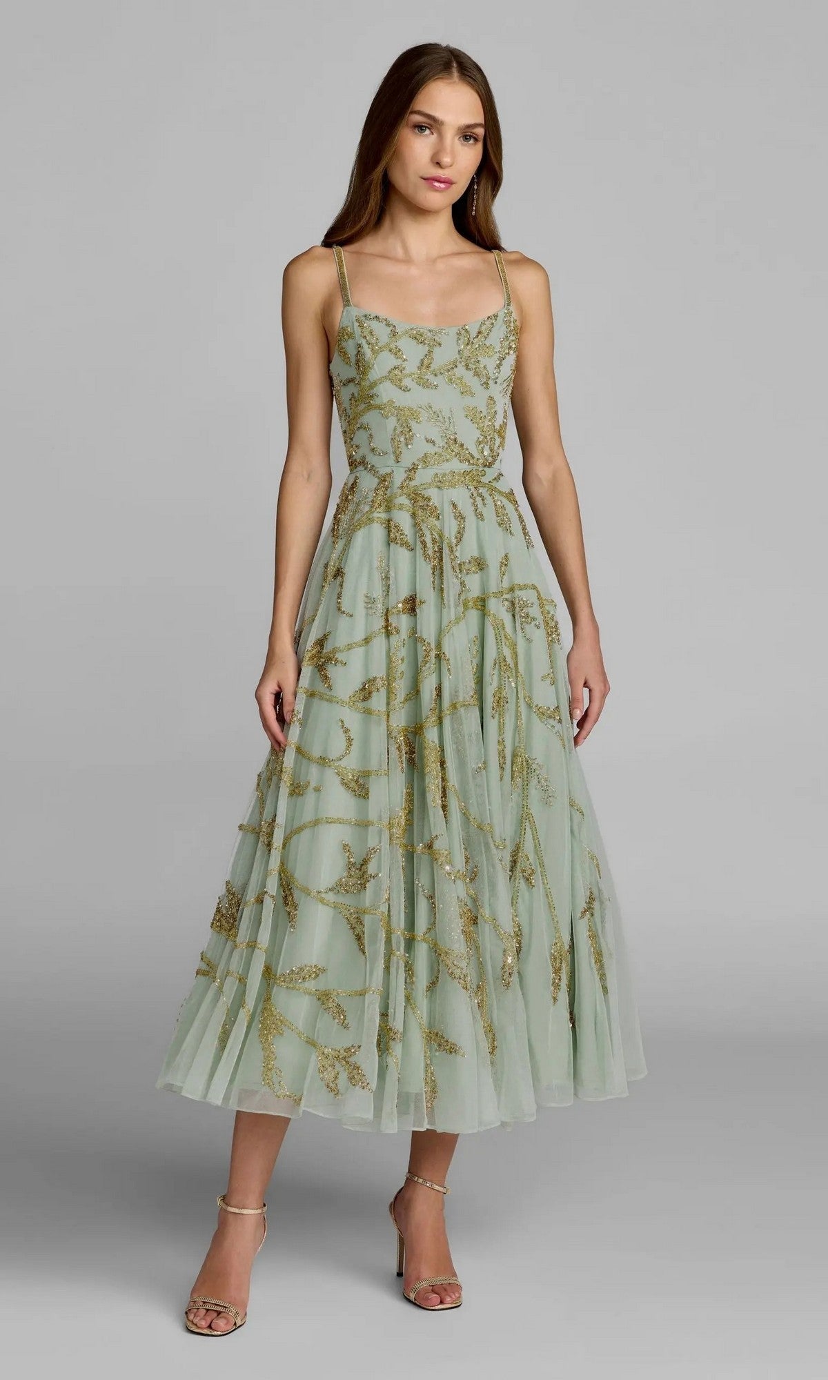 A woman stands on a gray background wearing the Tea-Length Party Dress: Mac Duggal 6255—a sleeveless, light green midi dress with gold embroidery—paired with strappy heels.