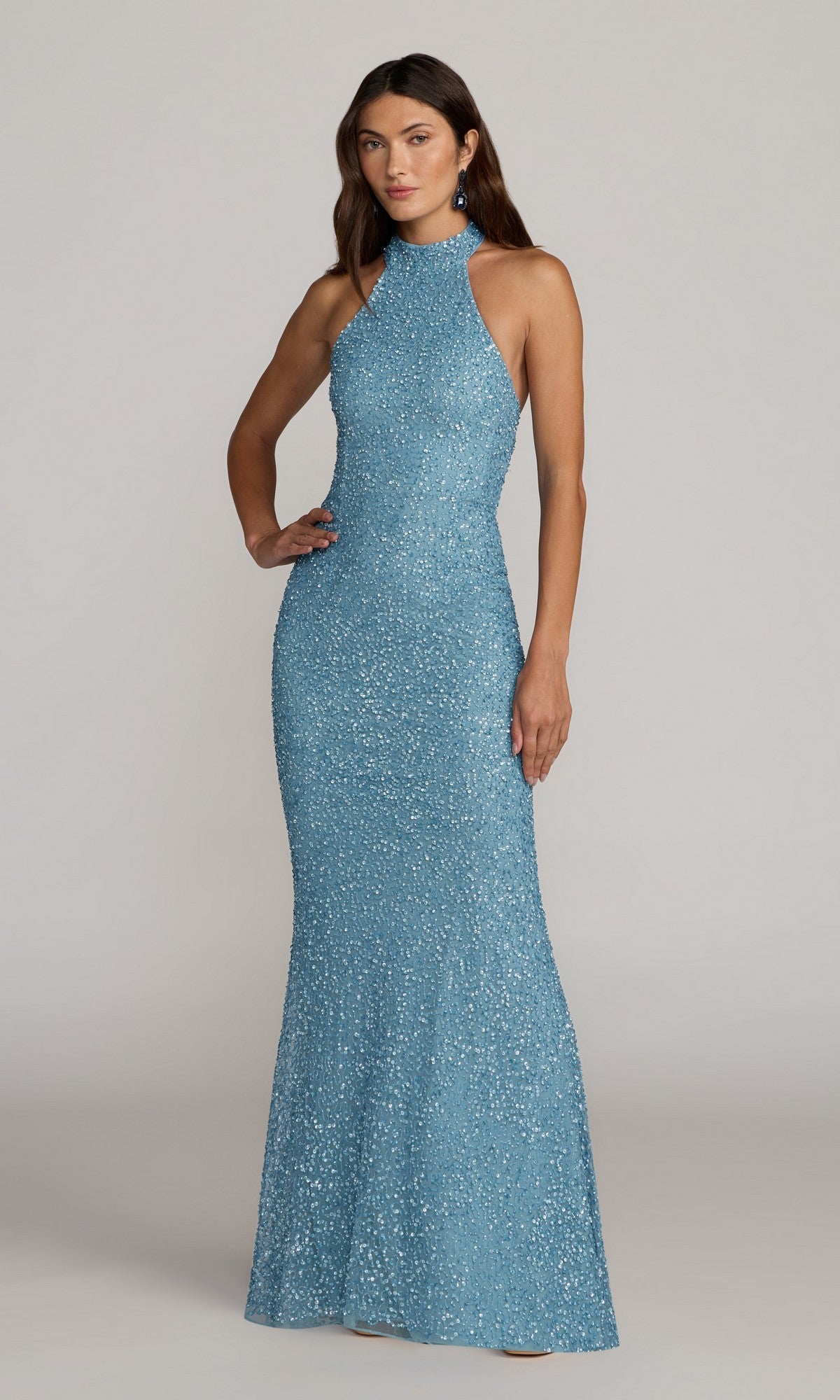 A woman stands with one hand on her hip against a neutral background, wearing the Mac Duggal 6259 Long Formal Dress in ice blue—a sleeveless, floor-length sequin gown with a high neckline.