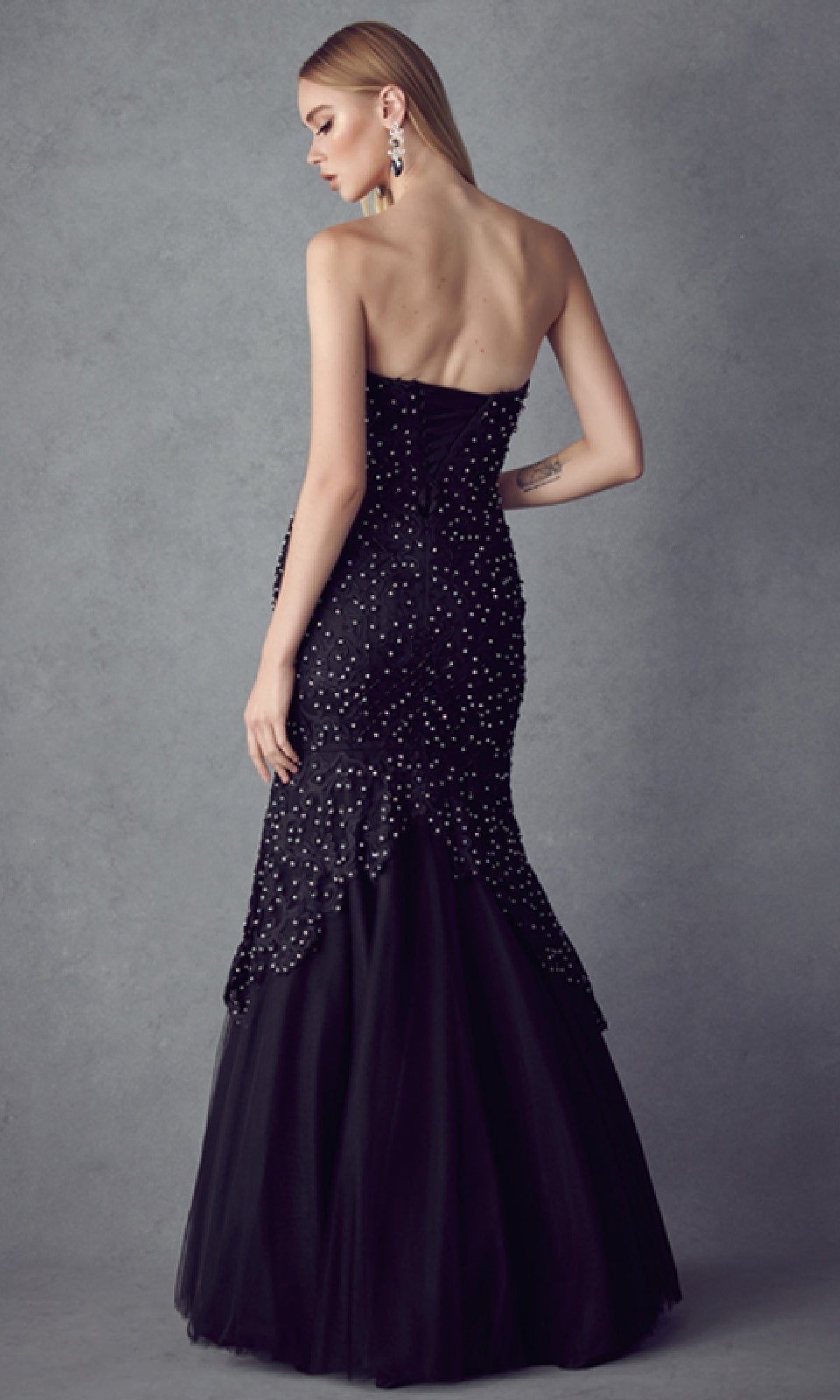 A woman wears the Long Mermaid Prom Dress: Juliet 644—black, strapless, floor-length with beaded details—posed against a gray background, facing away from the camera. This elegant gown is a stunning choice for formal events.