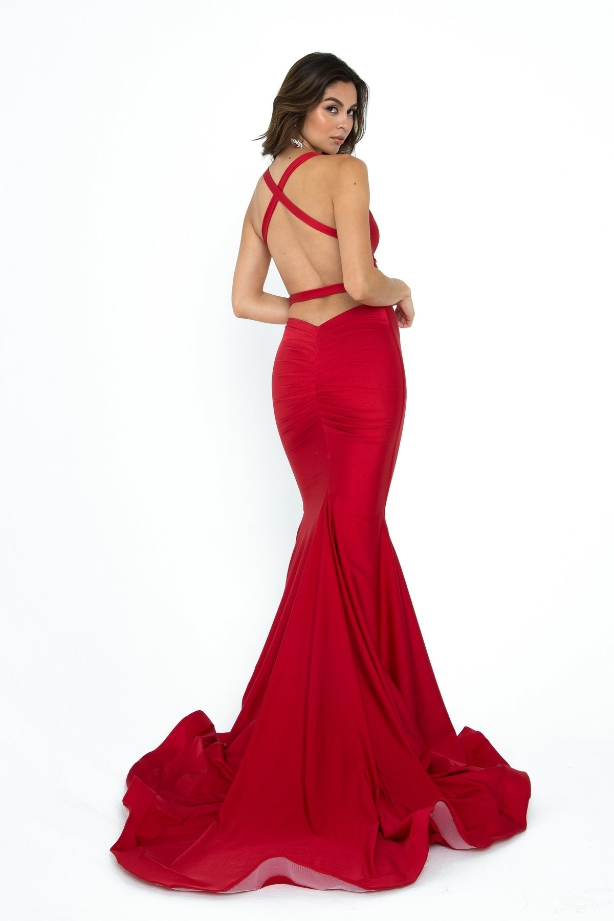 Woman wearing the Atria 6502H Open-Back Long Prom Dress—a fitted, floor-length red gown with a dramatic train and crisscross straps—posing against a plain white background.