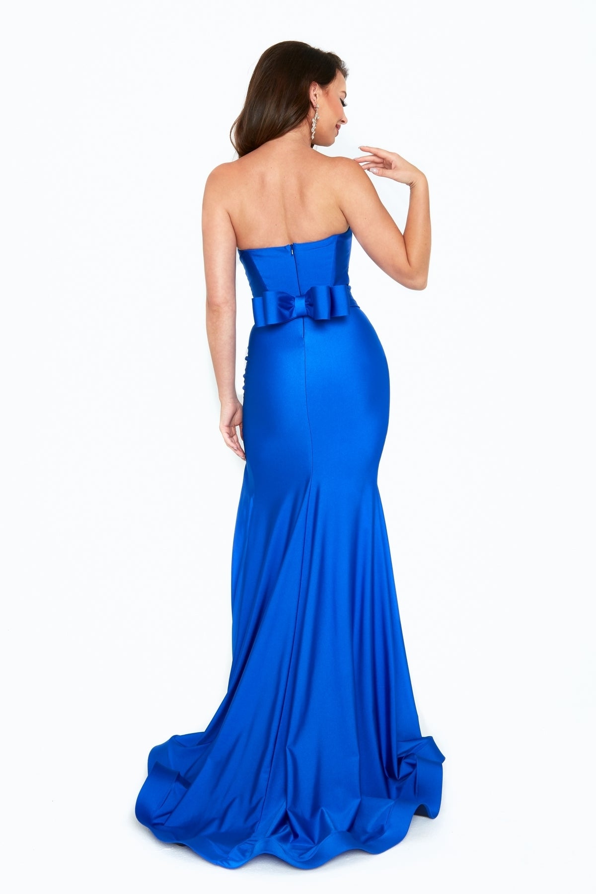 A woman in the Atria 6524H Strapless Long Prom Dress, floor-length and royal blue with a large back bow, stands facing away to highlight the gown's elegant back details.