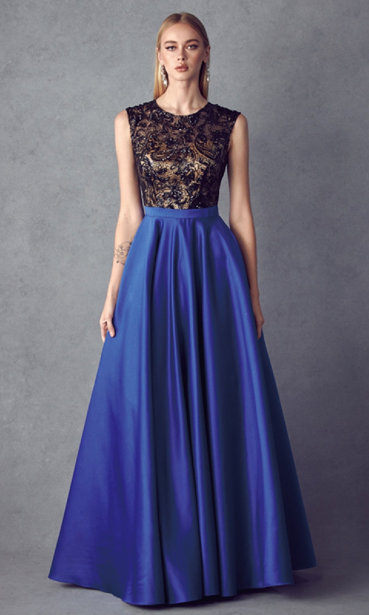 A woman wears the Cut-Out Long A-Line Prom Dress: Juliet 661, featuring an embellished black and gold bodice and a flowing royal blue skirt, as she stands against a plain gray background.